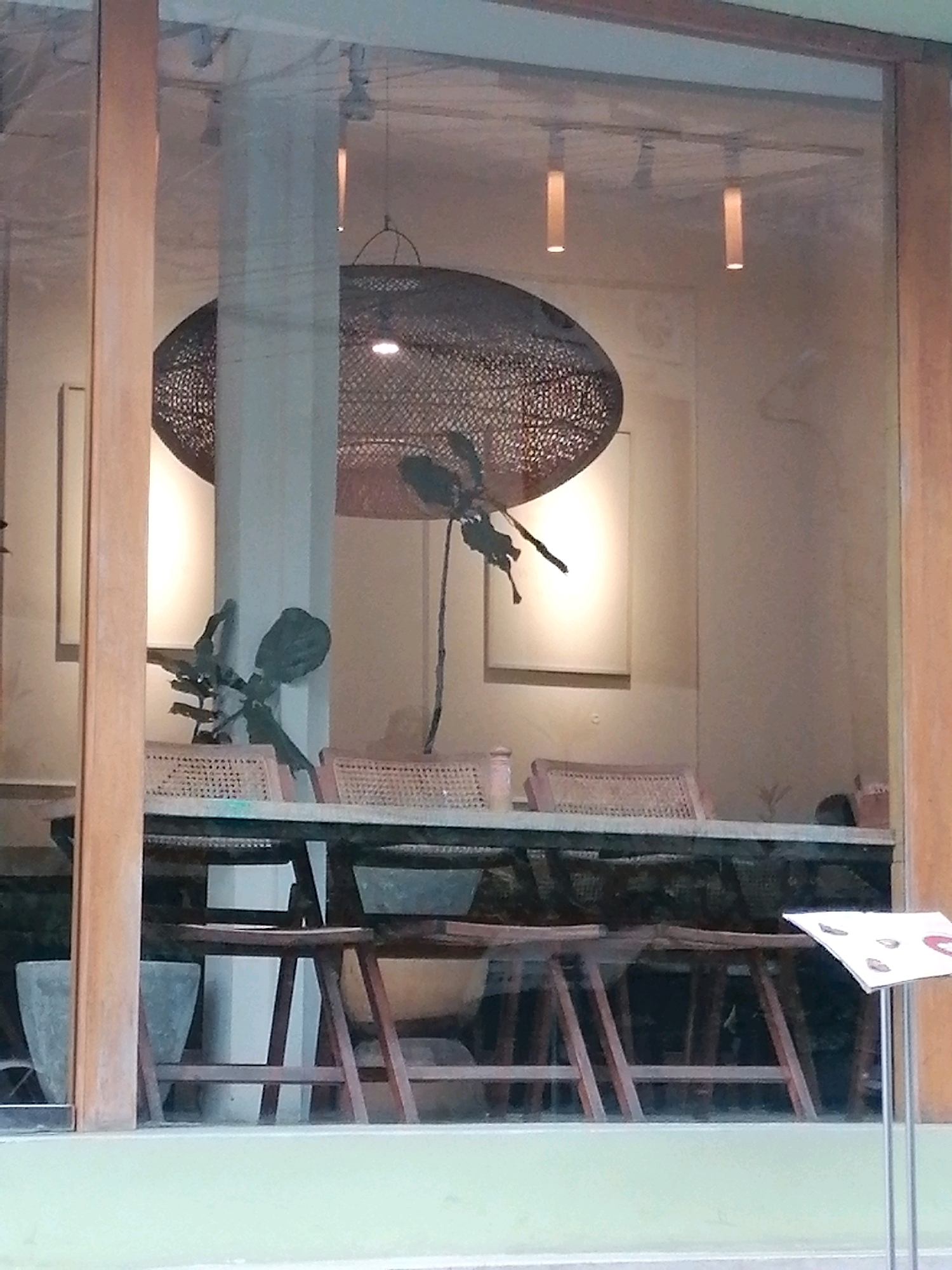 The image captures a view through a glass window into a brightly lit interior, possibly a cafe or restaurant, in Ubud District, Indonesia. The foreground features wooden window frames and a light green painted sill. Inside, a large, intricately woven lampshade hangs from the ceiling, illuminating the space. Two rectangular pendant lights with a warm glow are also visible. In the mid-ground, a long table is set with chairs featuring woven seats and wooden frames. A decorative plant with dark, sculptural foliage is positioned near a white pillar. Two framed artworks hang on the back wall. The overall aesthetic appears modern with natural elements, suggesting a contemporary design.