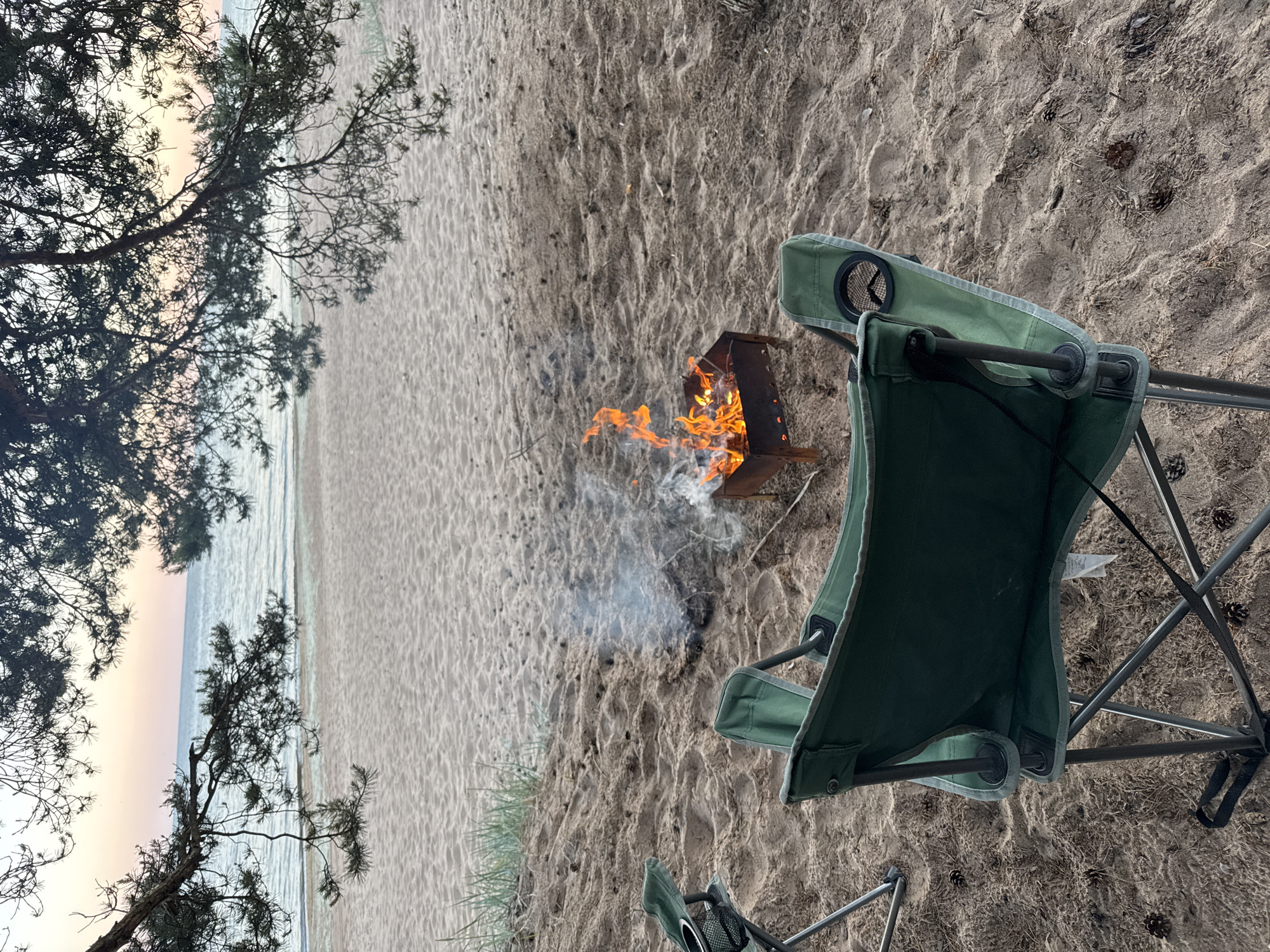 An outdoor scene at dusk on a sandy beach shows a small metal barbecue with an active fire, producing smoke. Two green folding chairs are positioned on the sand in front of the barbecue, with one chair closest to the viewer. Pine branches hang over the scene from the top. The water of a calm sea is visible in the background, with a soft gradient of pink and pale blue in the sky above it. The beach is sparsely scattered with pinecones and dry grass. The location is the beach of Zelenaya Roshcha, Russia.