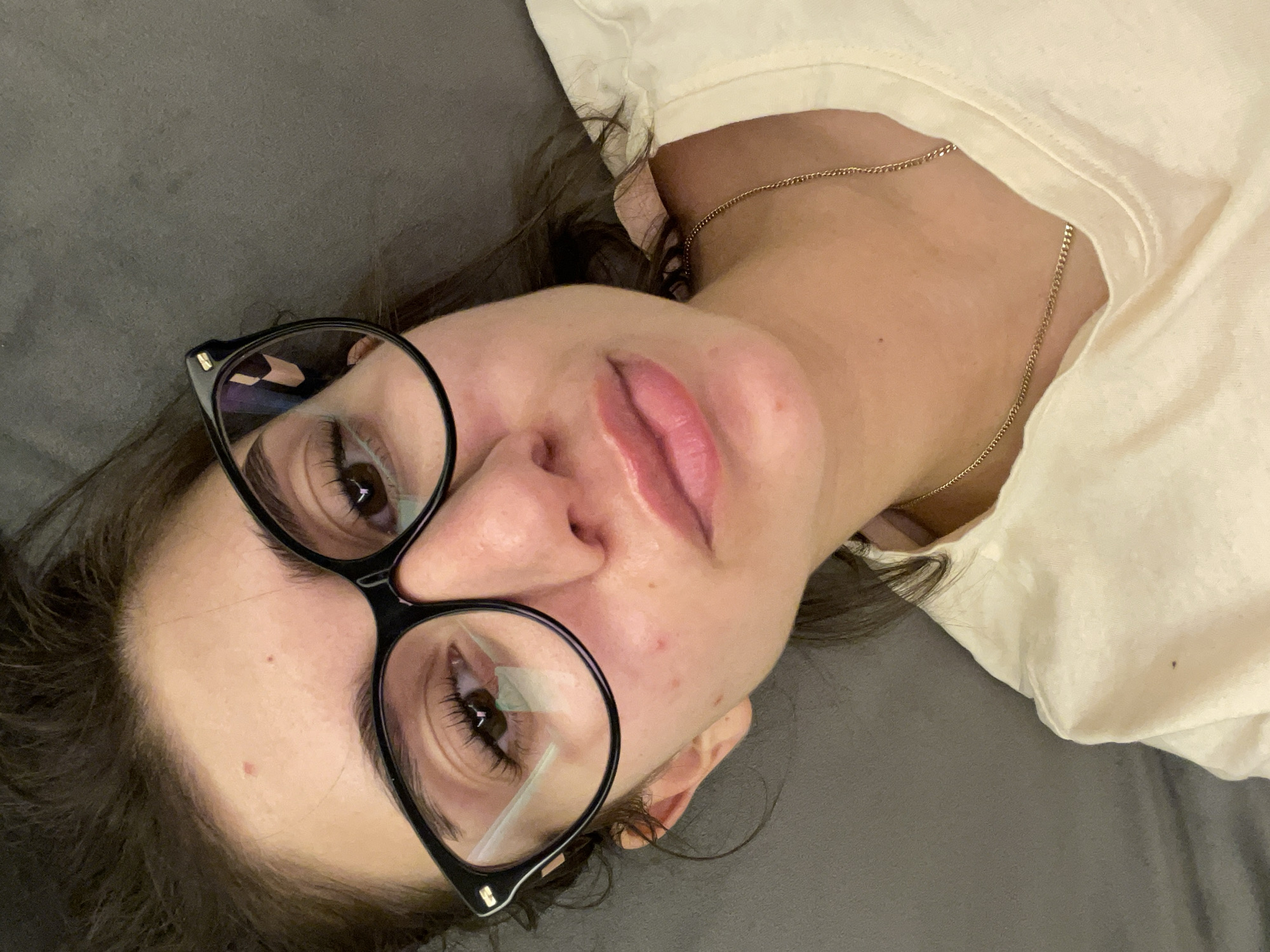 A woman is lying on a gray surface, likely a couch or bed, wearing black-framed glasses and a light-colored t-shirt. A gold chain necklace is visible around her neck. The location cannot be determined from the image.