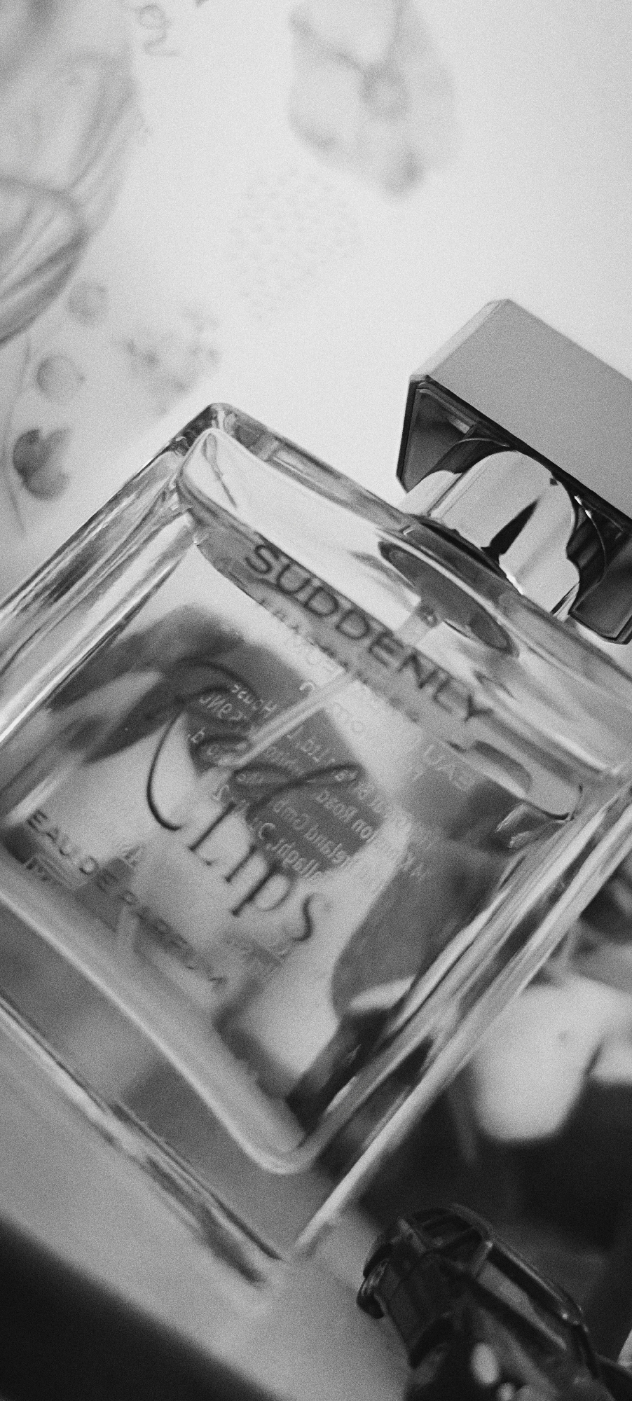 This is a black and white, close-up, angled shot of a perfume bottle with a silver cap. The bottle is clear glass, and the word "SUDDENLY" is embossed on its side. Below "SUDDENLY," the word "LIPS" is also visible, along with what appears to be "EAU DE PARFUM" on the lower left. The bottle rests on a surface with a blurred, textured background, possibly a piece of fabric or paper with floral or abstract patterns. The lighting creates reflections and highlights on the glass. The image does not provide any information or visual cues to indicate a specific location in Winnenden, Germany.