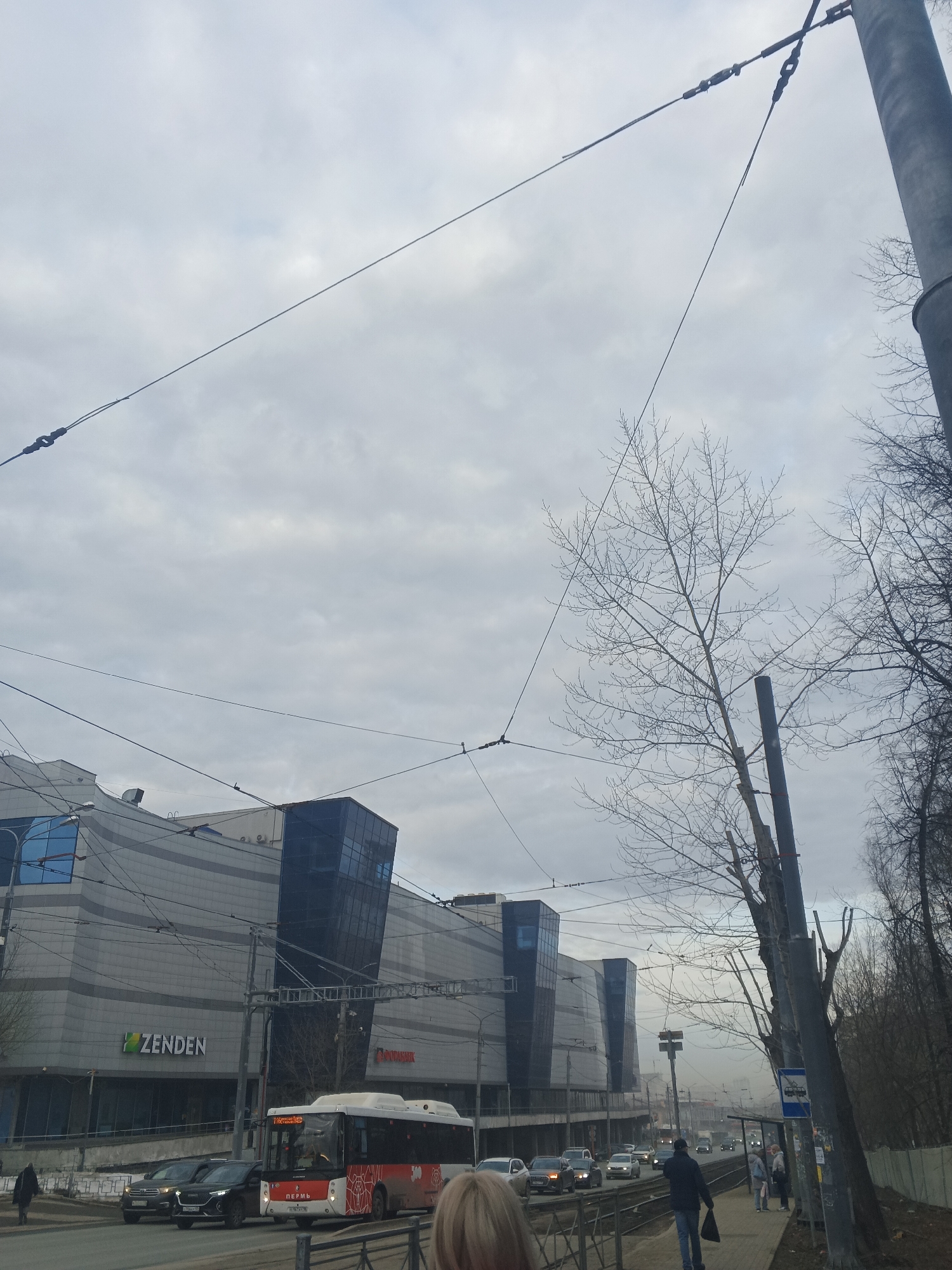 The image depicts a street scene in Perm, Russia, likely taken during the colder months due to the bare trees and overcast sky. A modern, multi-story building with a prominent "Zenden" sign is visible on the left. Along the street, there are several cars and a red and white bus marked with "Пермь" (Perm) in Cyrillic. A bus stop sign is located next to some bare trees. A few individuals are walking along the sidewalk, and overhead tram lines are suspended. The sky is heavily overcast, suggesting a cloudy day.