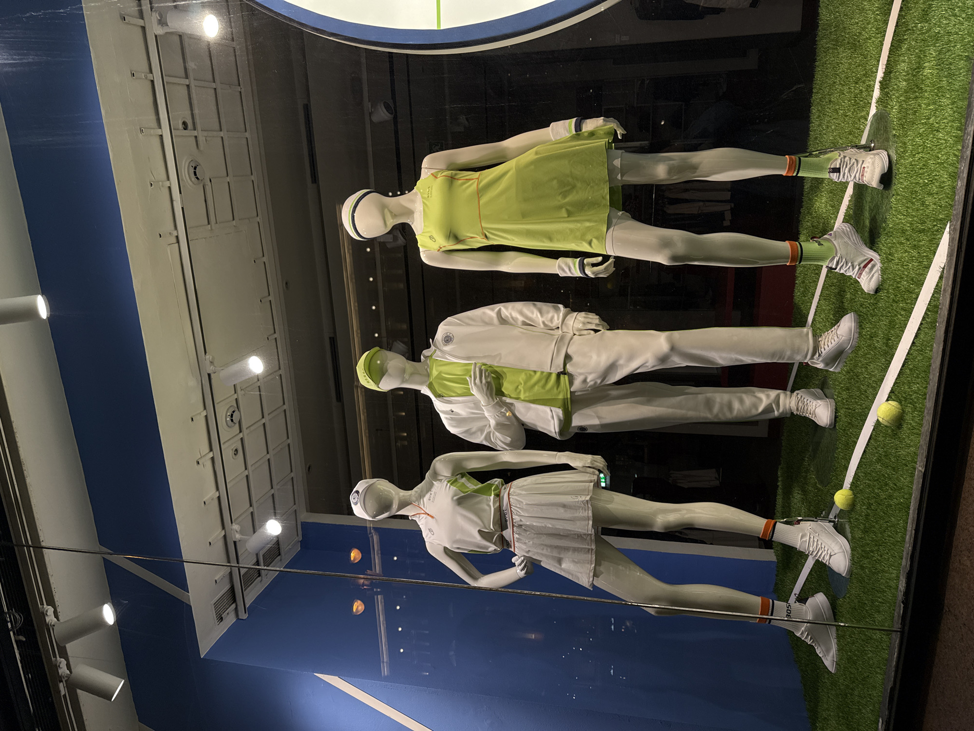The image shows a window display of three mannequins dressed in tennis apparel. The mannequins are posed on a green surface meant to simulate a tennis court. There are tennis balls present. The mannequin on the top is wearing a lime green tennis dress and matching accessories. The middle mannequin is wearing a white jacket and white pants with lime green accents. The bottom mannequin is wearing a white tennis skirt and matching top with orange accents. The backdrop of the window display has blue and white painted elements that appear to mimic a tennis court. Reflections in the glass indicate the presence of lighting fixtures and a grid-like ceiling structure. There are no people visible in the background beyond the reflections. The location is Moscow, Russia.