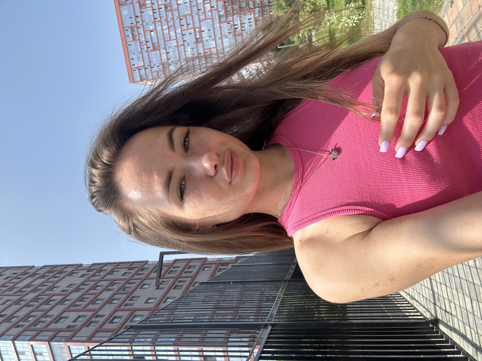 An outdoor, eye-level, medium shot captures a young, light-skinned woman with long, light brown hair against a background of a bright, clear blue sky. She is wearing a pink tank top and a silver necklace. Her right arm is bent, with her hand resting on her chest. In the background, two modern, red and beige apartment buildings stand tall, with a black metal fence in the foreground. The sunlight casts shadows on the paved ground.