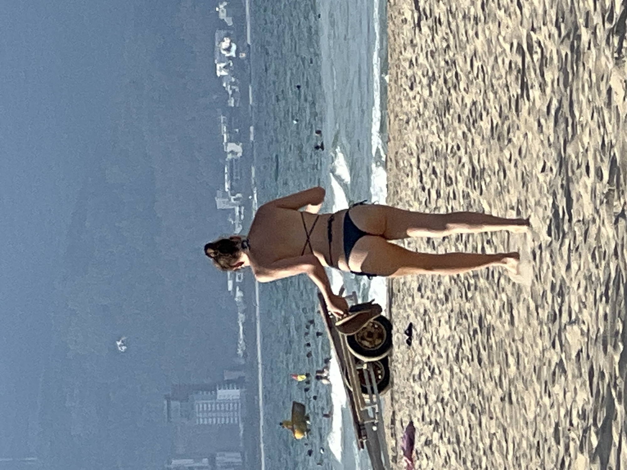 In Da Nang, Vietnam, a woman walks on a sandy beach toward the ocean. She is wearing a dark bikini and carrying a pair of brown sandals in her right hand. Behind her, the ocean has waves, and several people are swimming. In the distance, there is a yellow boat, a boat trailer with two wheels on the sand, and buildings on a hazy coastline. The sky is bright and clear.