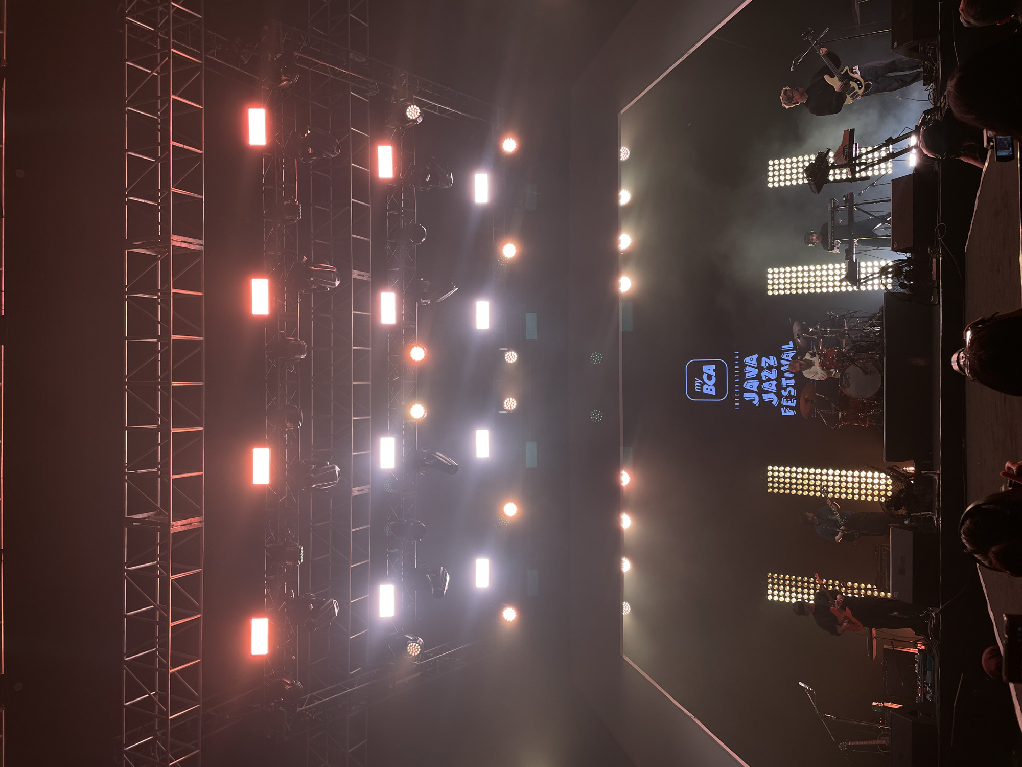 The image depicts a concert stage at the My BCA International Java Jazz Festival, likely in Salembaran, Indonesia. The stage is lit with numerous stage lights arranged on metal rigging structures to the left. A large screen displays the festival logo. Musicians are performing on stage, including guitarists, keyboardists, and a drummer. Spectators are visible in the foreground.