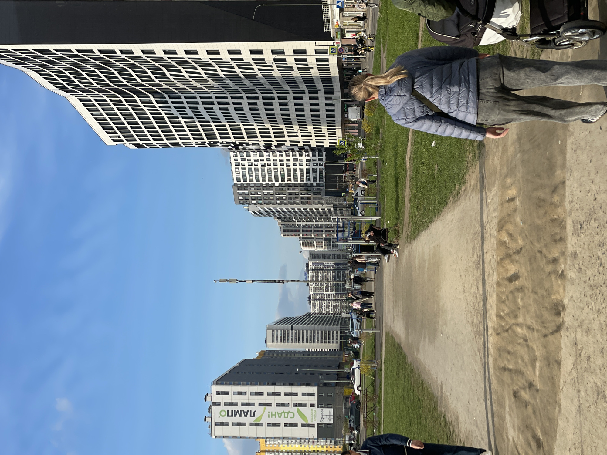 Here is a description of the image:
**Scene:** An urban park setting in Unknown, Russia, featuring modern high-rise buildings in the background. The sky is clear and blue.
**People:** A woman in a blue jacket and gray pants is walking with a stroller on a dirt path. There are other people, including children, scattered along the path and on benches in the park. Some are sitting on benches, while others are strolling.
**Objects:** High-rise buildings with a modern design dominate the background. There is green grass along the path and trees providing some shade. Other objects include a stroller, benches, and a tall metal structure. One building features a prominent advertisement.
**Actions:** People are walking, sitting, and generally enjoying the park. The woman is pushing a stroller along the path.
**Interactions:** People are interacting with each other in the park, such as walking together or sitting and talking. The woman is focused on her stroller.
**Environment:** The scene is well-lit with sunlight. The park appears clean and well-maintained, with a mix of open space and landscaped areas. The modern architecture of the buildings gives the environment a contemporary feel.