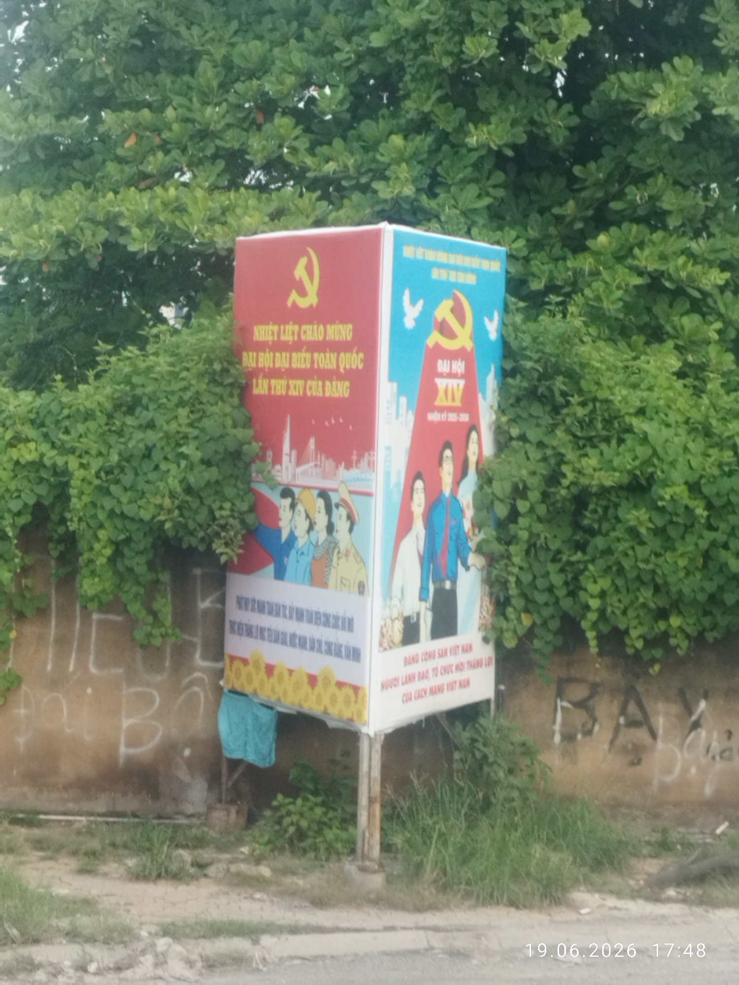 This image captures a roadside scene in Ho Chi Minh City, Vietnam, characterized by a vibrant propaganda banner displayed on a metal pole. The banner, divided into two sections, features prominent communist symbols, including a yellow hammer and sickle on a red background and on a blue background. The red section of the banner includes Vietnamese text, which translates to "Warmly welcome the 14th National Congress of the Party" and depicts stylized illustrations of people and a city skyline with a bridge. The blue section also contains Vietnamese text and illustrations, with a focus on the "XIV Congress" and the years "2025-2030."
The banner is positioned in front of a weathered, light brown wall adorned with graffiti, including the letters "HE" and "Đai Bô" in white. To the right of the banner, more graffiti is visible, with the letters "BAY" and illegible characters in black and white. Lush green foliage, likely bushes and trees, partially obscures the wall and the banner, creating a contrast between the man-made structure and the natural environment. In the foreground, a patch of grass and some scattered debris indicate a slightly unkempt area. A turquoise piece of fabric hangs from a makeshift structure behind the banner, adding a touch of everyday life to the scene. The lower right corner of the image displays a timestamp: "19.06.2026 17:48". The overall impression is one of a typical urban landscape in Vietnam, where political messaging, graffiti, and the surrounding greenery coexist.
