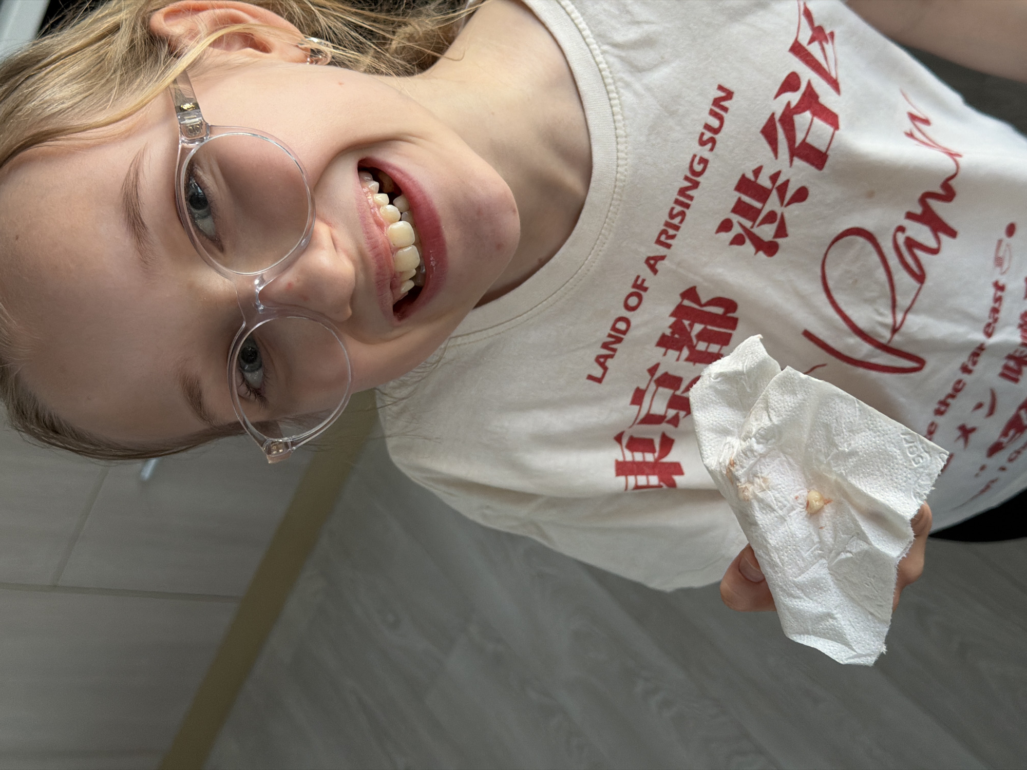 A young girl with blonde hair pulled back and wearing clear framed glasses is smiling broadly, showing her teeth. She is holding a crumpled tissue with a small speck of what appears to be food. She's wearing a white t-shirt with red Japanese writing, including the English phrase "LAND OF A RISING SUN" and "Ramen" on it. The background appears to be a light gray wall with a wood-like pattern, the scene is indoors. The location of Dreiliņi, Latvia, cannot be confirmed based on visual information.