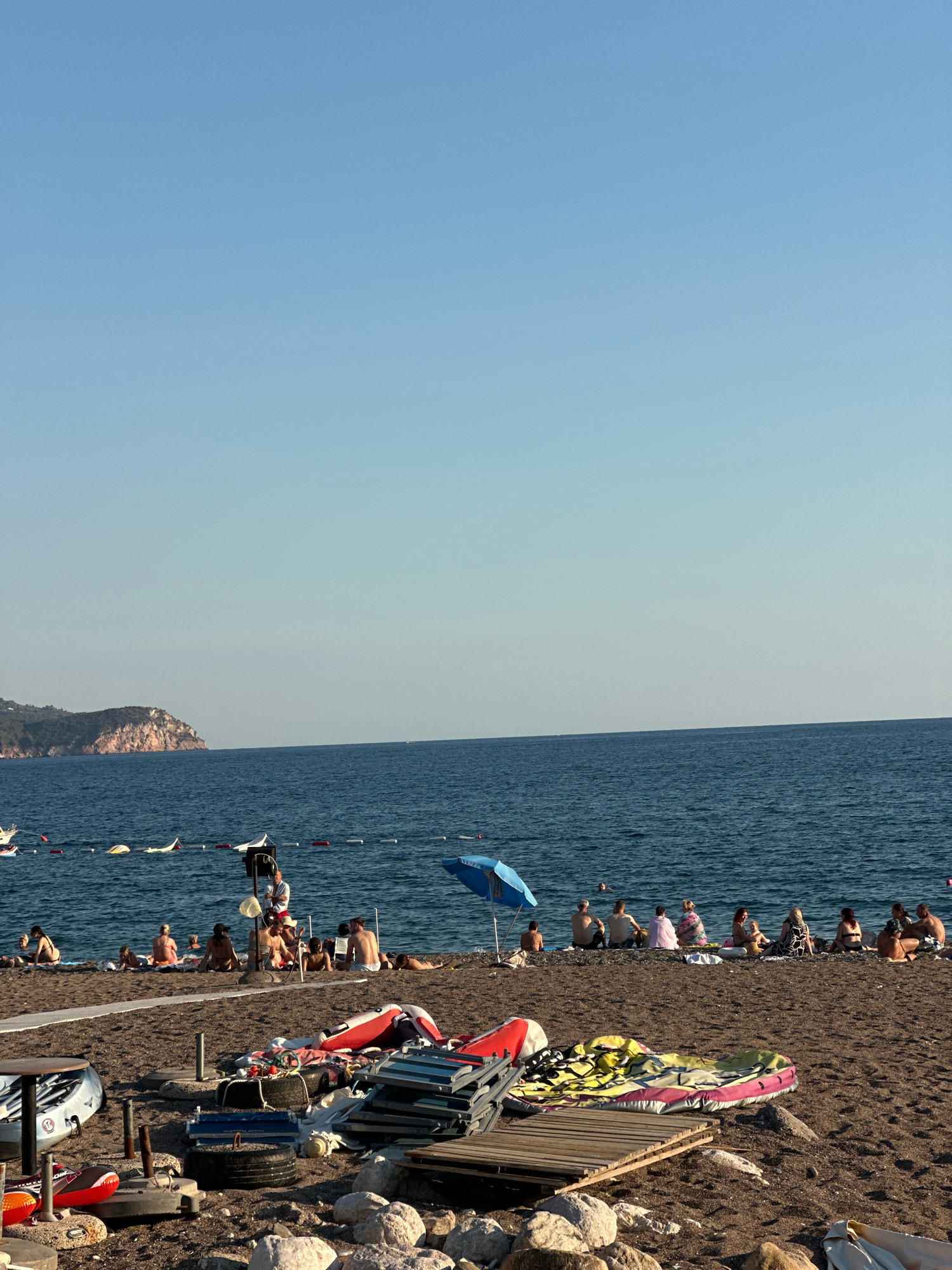The image depicts a sunny day at a pebble beach in the city of Boreti, Montenegro. The sea is calm and a clear blue, with a distant headland on the left. In the foreground, various beach equipment is piled up, including deflated inflatable boats, chairs, tires, and wooden planks. A person is seated on a chair near a white device with a microphone on the beach. A group of people are sitting on the beach facing the sea, some are sunbathing and others are conversing. A blue umbrella provides shade for some of the beachgoers. The scene is serene, with people enjoying a day at the seaside.