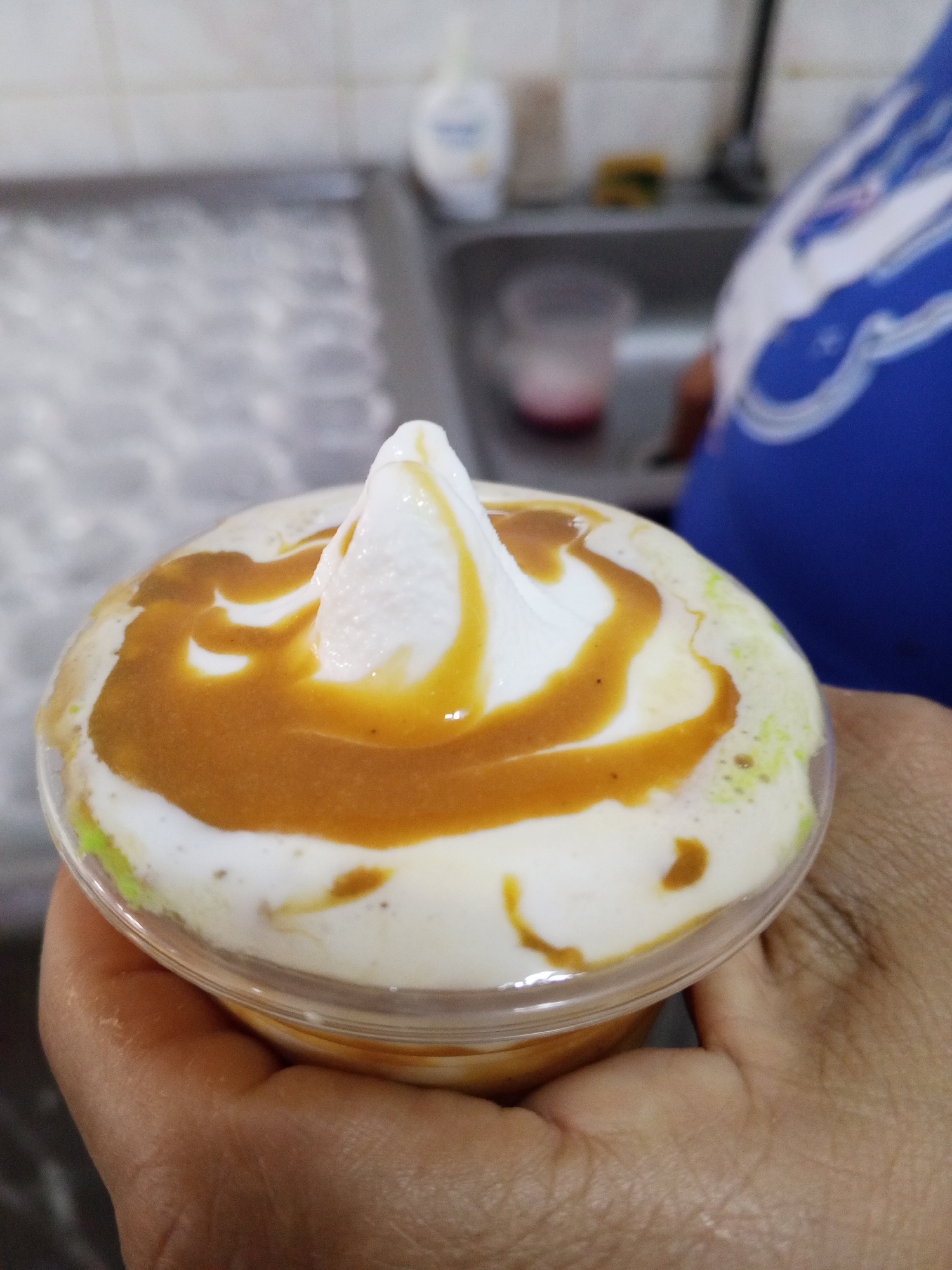 A close-up shot depicts a hand holding a clear plastic cup filled with a dessert. The dessert consists of a base layer, topped with a white creamy substance, likely soft-serve ice cream, and drizzled with a brown caramel-like sauce. A swirl of the creamy substance rises to a peak in the center. The hand holding the cup appears to belong to a person with tan skin. The background shows a blurry countertop with some objects, including a container of lotion and what appears to be a sink, and a person wearing a blue shirt with white detailing. The location is Tinaquillo, Venezuela.