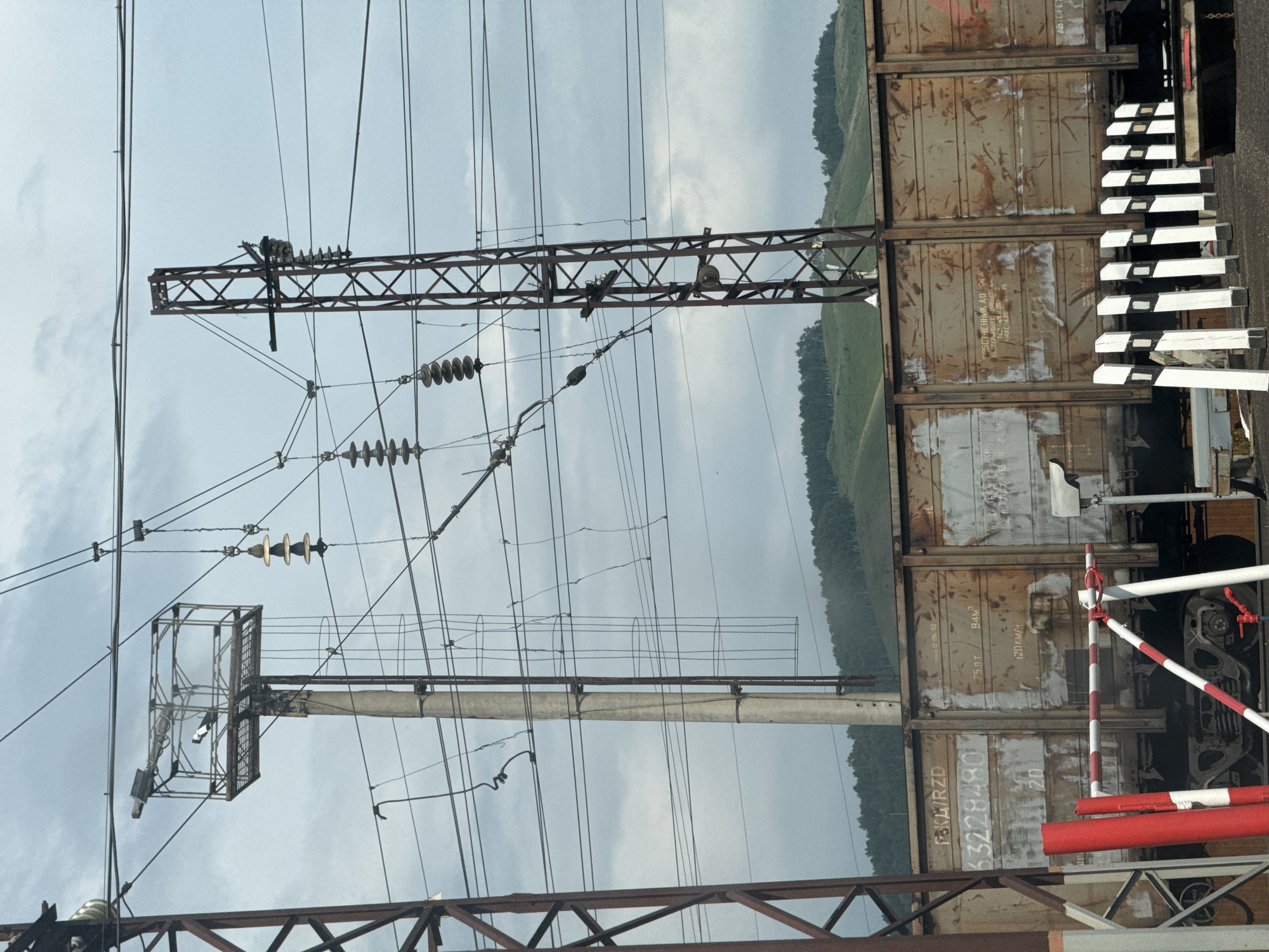 This image shows a train at a railway crossing on a cloudy day in Zykova, Russia. The train cars are weathered and rusty, with Cyrillic text and numbers visible on their sides. A railway crossing gate with red and white stripes is positioned in the foreground. Overhead, a complex network of electrical wires and insulators, supported by concrete and metal poles, spans across the frame. In the background, rolling hills covered in lush green trees are visible under a grey, overcast sky. The scene depicts typical infrastructure and landscape of a rural railway in Russia.