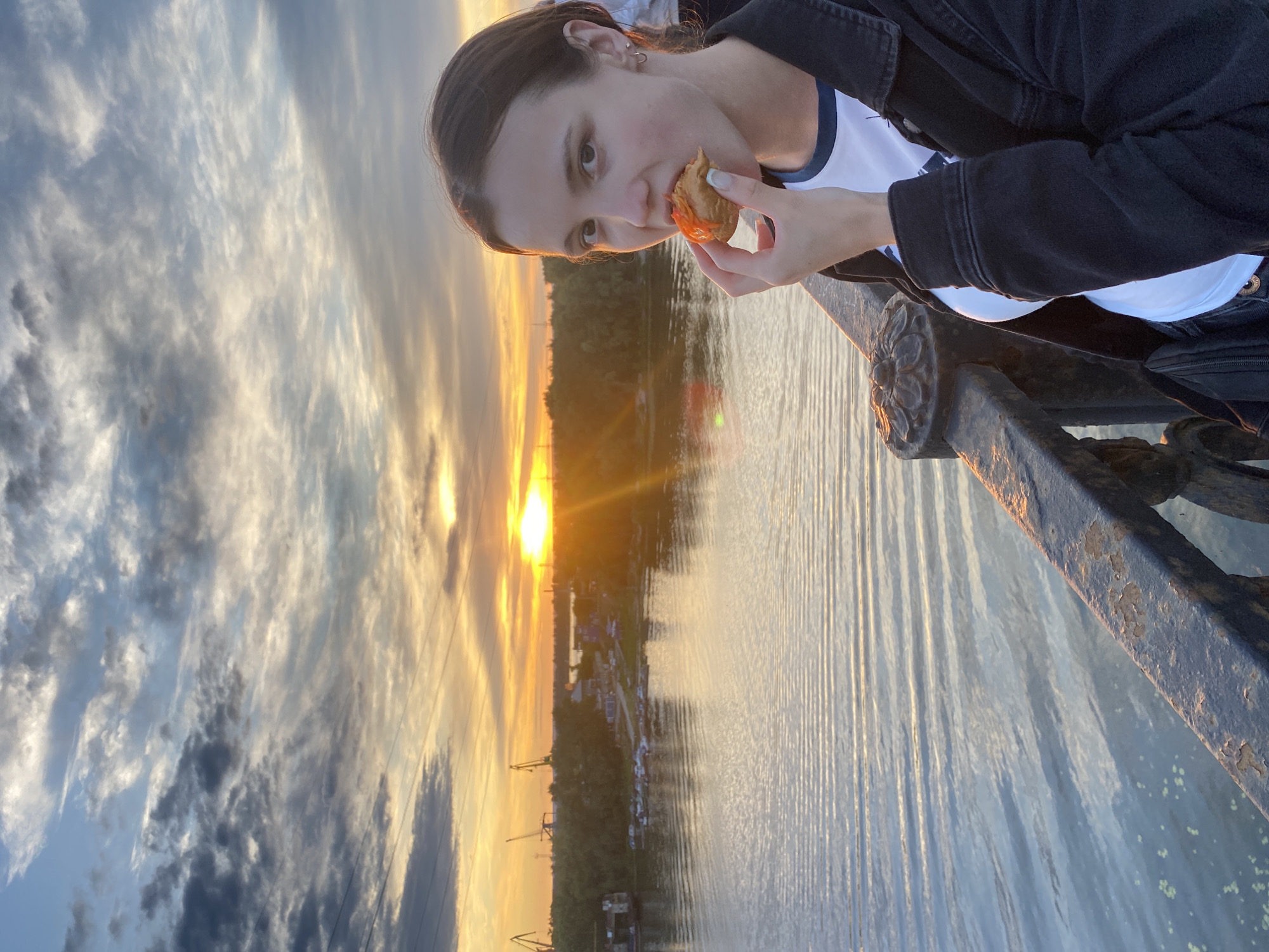 A young woman is eating a pastry with orange filling while standing on a bridge overlooking a river at sunset. The sky is filled with dramatic clouds illuminated by the setting sun, casting a warm glow over the scene. The river is calm, with gentle ripples reflecting the sky. On the far bank, trees line the shore, and industrial cranes are visible in the distance, suggesting an urban or industrial setting. The woman is wearing a dark jacket over a white t-shirt and has dark hair. The bridge railing in the foreground is made of dark metal with decorative elements. The overall impression is one of a peaceful evening scene, possibly in Novokuybyshevsk, Russia, given the presence of industrial infrastructure and the general landscape.
