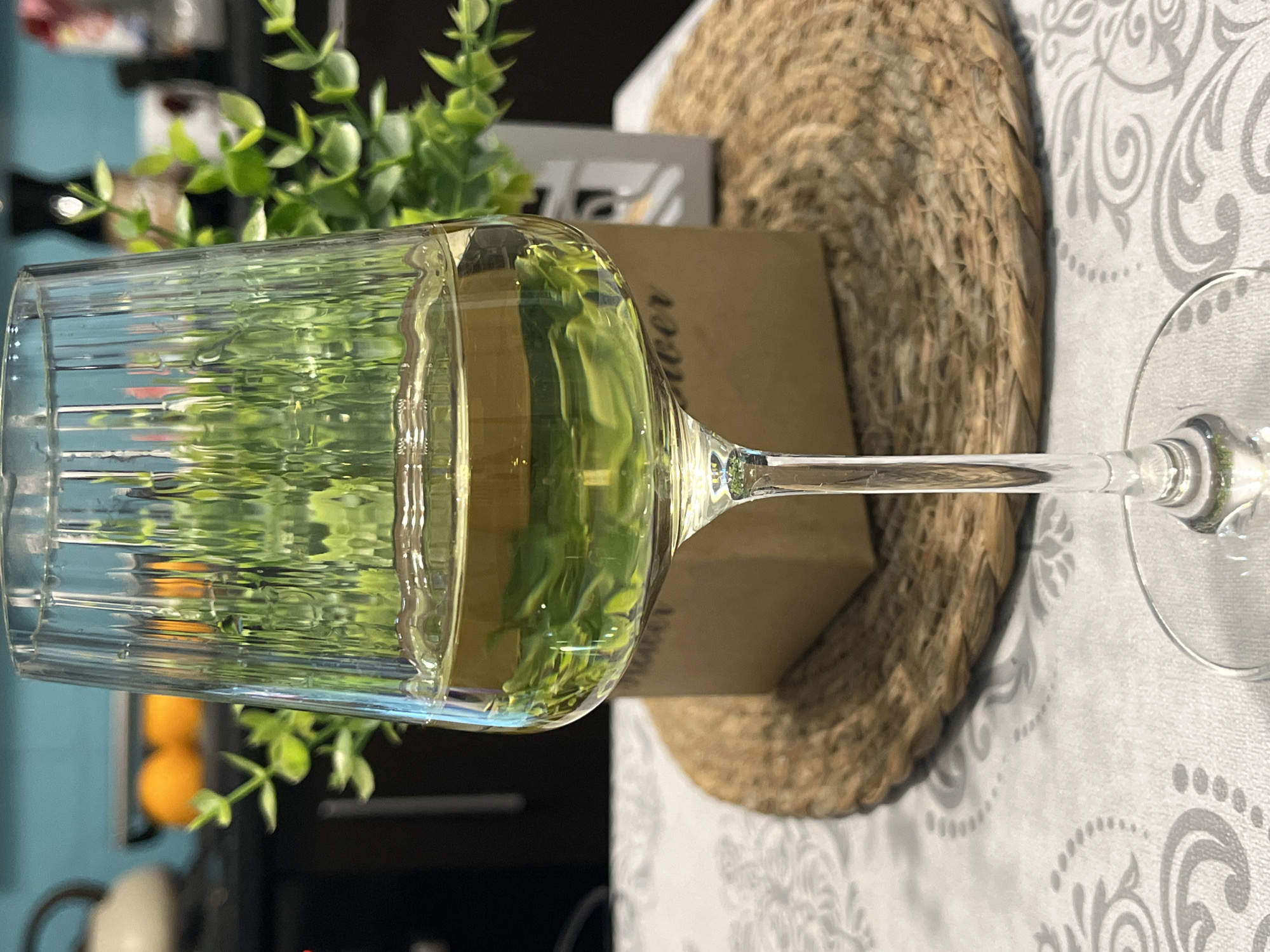 A horizontal medium shot depicts a tall, ridged glass filled with a pale yellow liquid, likely wine, set against a blurred background. The glass sits on a patterned white tablecloth next to a circular, woven placemat. Behind the glass, out of focus, is a potted green plant and a rectangular object with text. The scene appears to be indoors, in a domestic setting. Due to lack of information I cannot infer the location to be Tsentral'noiusad'byopytnoistantsiivniimk, Russia.