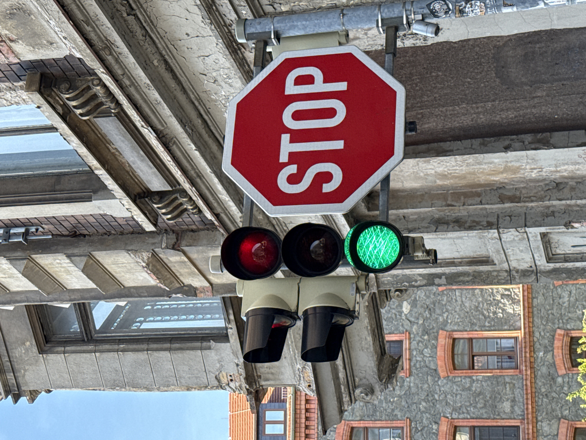 The image depicts a street scene in Görlitz, Germany, focusing on a traffic light and a stop sign. The traffic light shows a red light at the top and a green light at the bottom, with the middle light off. The stop sign is octagonal, red with a white border, and displays the word "STOP" in white capital letters. The traffic light and stop sign are mounted on a metal pole. In the background, there is an old building with stone and brick facades, featuring windows with dark frames. Another building with a red tiled roof and dormer windows is visible to the left. The sky is clear and blue. The image is taken from a low angle, looking up at the traffic control devices and buildings.