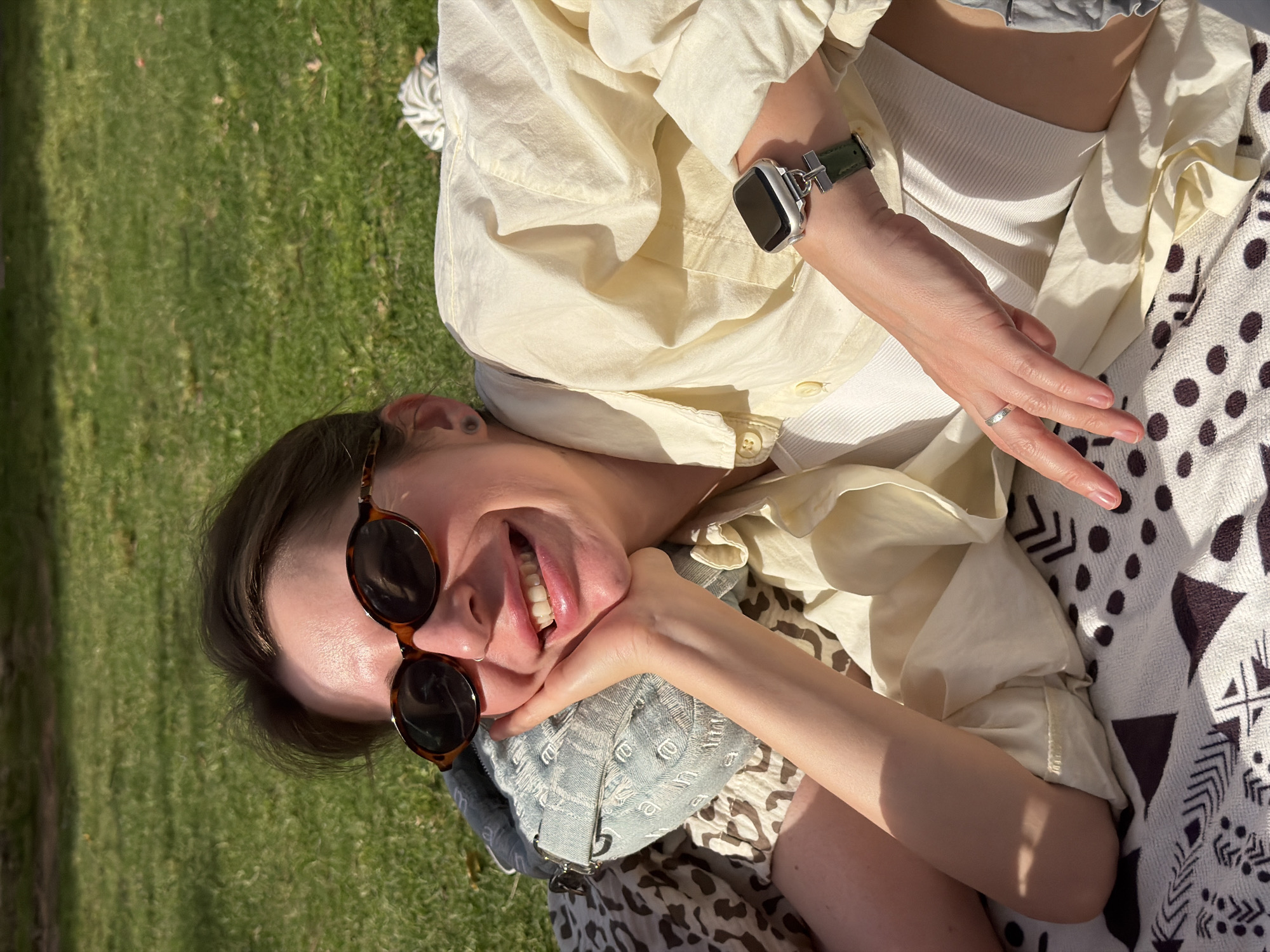 A young woman is lying on a patterned blanket outdoors on a sunny day, possibly in a park. She is wearing sunglasses and an unbuttoned beige shirt over a white tank top. She has a smartwatch on her left wrist and a ring on her left hand. The background includes green grass and a play of light and shadows. The location is not discernible as Tel-Aviv without additional landmarks or cues.