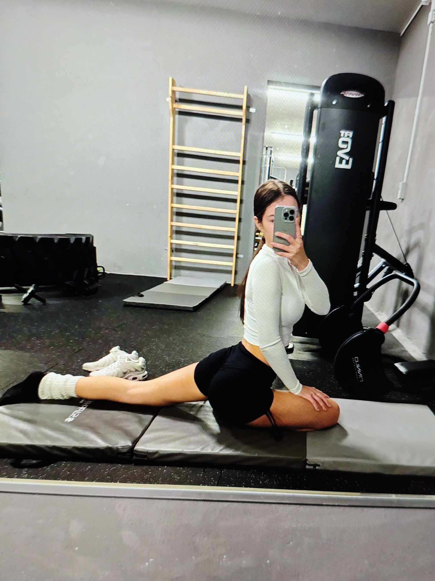 A woman is performing a stretching exercise on a yoga mat in a gym. She is wearing a white long-sleeved crop top, black shorts, white socks, and white athletic shoes. Her right leg is extended forward, with her foot pointed, while her left leg is bent backward, with her knee on the mat. She is holding a smartphone in her right hand, taking a selfie.
The gym has a gray wall and a black rubber floor. In the background, there is a wooden wall rack with horizontal bars, a black weight machine, and other gym equipment. The lighting in the gym appears to be artificial. The scene is captured from a slightly low angle, with a mirror reflecting the woman and her surroundings.
The image is set in a gym located in Baku City, Azerbaijan.