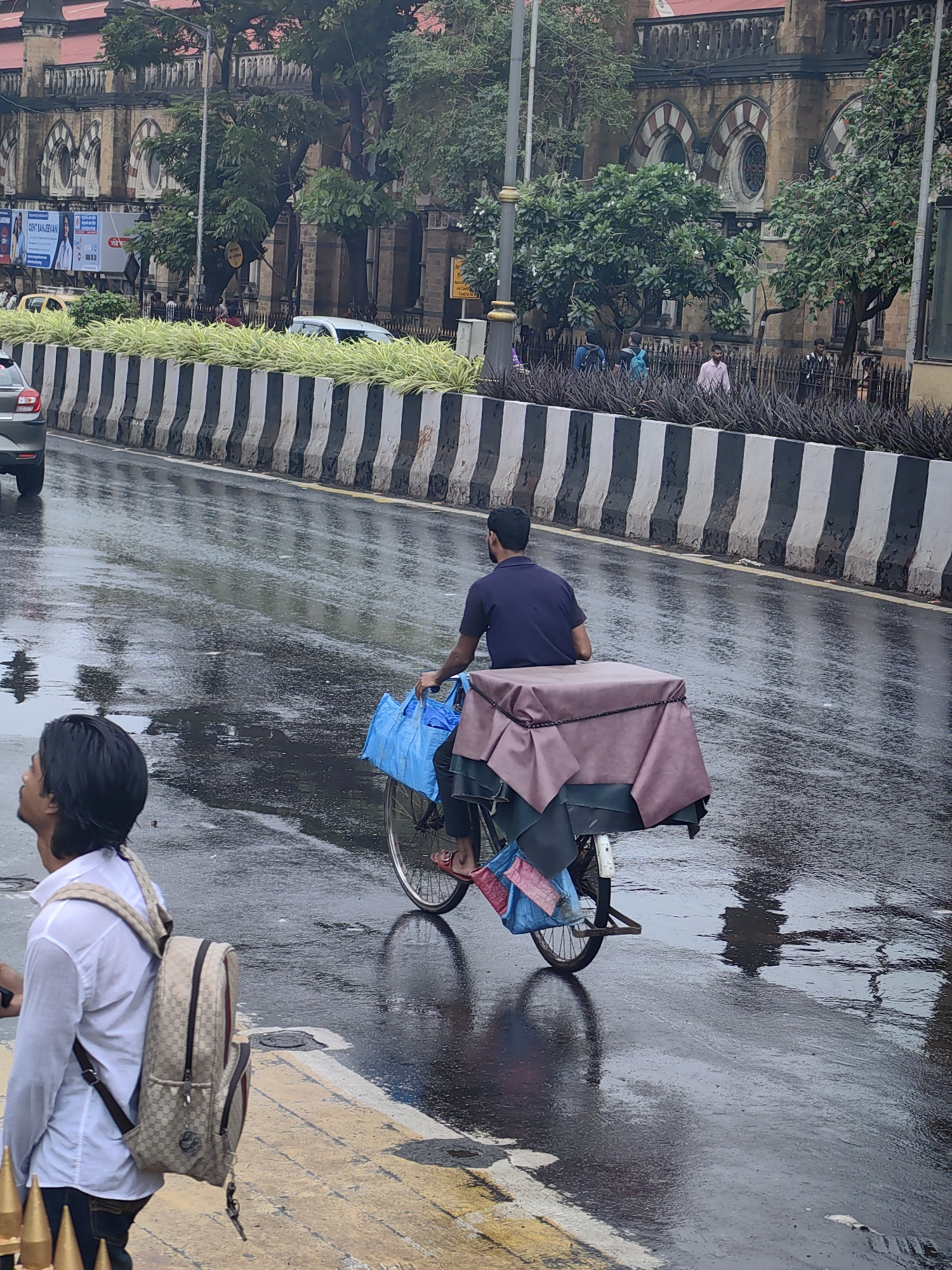 The image depicts a street scene in Mumbai, India, likely during or after a rain shower, as the asphalt is wet and reflective. In the foreground, a young man wearing a white collared shirt and dark denim jeans is seen from the back, looking up and to the left. He is carrying a patterned backpack with leather accents. To his right, a man on a bicycle is riding away from the viewer. He wears a dark blue t-shirt and is transporting a large, covered load on the rear of his bicycle. The load is covered with a pinkish-brown tarp secured with a rope. A bright blue bag is also attached to the bicycle.
The background features a large, ornate building with arched windows, characteristic of colonial-era architecture, possibly a government building or a historic landmark in Mumbai. Trees line the sidewalk and the street. A dividing barrier with black and white stripes separates the road from the sidewalk. Several cars are visible on the wet road. The overall atmosphere is urban and busy, with evidence of recent rainfall.