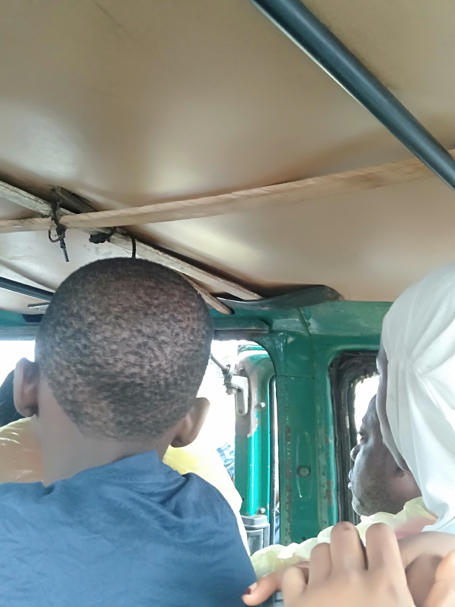 Here is a description of the image:
The photograph depicts the interior of a vehicle, possibly an auto rickshaw, transporting at least two individuals. A child with closely cropped hair wears a blue shirt and is viewed from the back. To the right, an adult wears a white head covering, and their profile is visible. The vehicle's frame is painted green, and the ceiling has a light color. A metal bar runs across the top of the frame. The setting is presumably within an urban area.