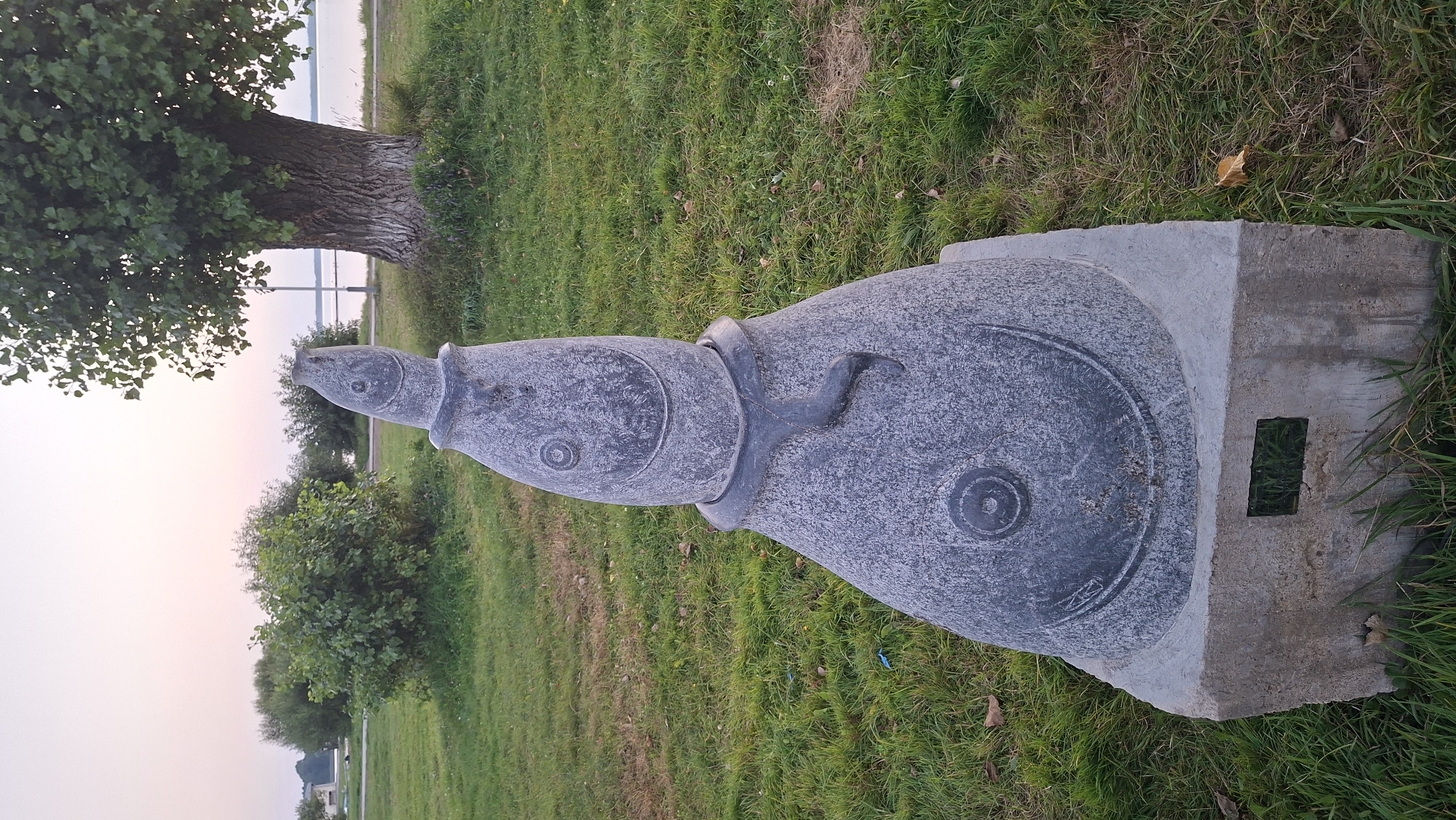 A stone sculpture resembling a stylized fish stands on a concrete pedestal in a grassy park area. The sculpture is gray and has circular designs for eyes and a mouth. A dark, snake-like element is wrapped around its neck. In the background, there are green bushes, a large tree with textured bark, and a body of water with a distant shoreline. The sky is a soft gradient of pink and pale blue, suggesting dawn or dusk. The scene appears to be outdoors in a public space, likely a park or waterfront area in Myadzyel, Belarus.