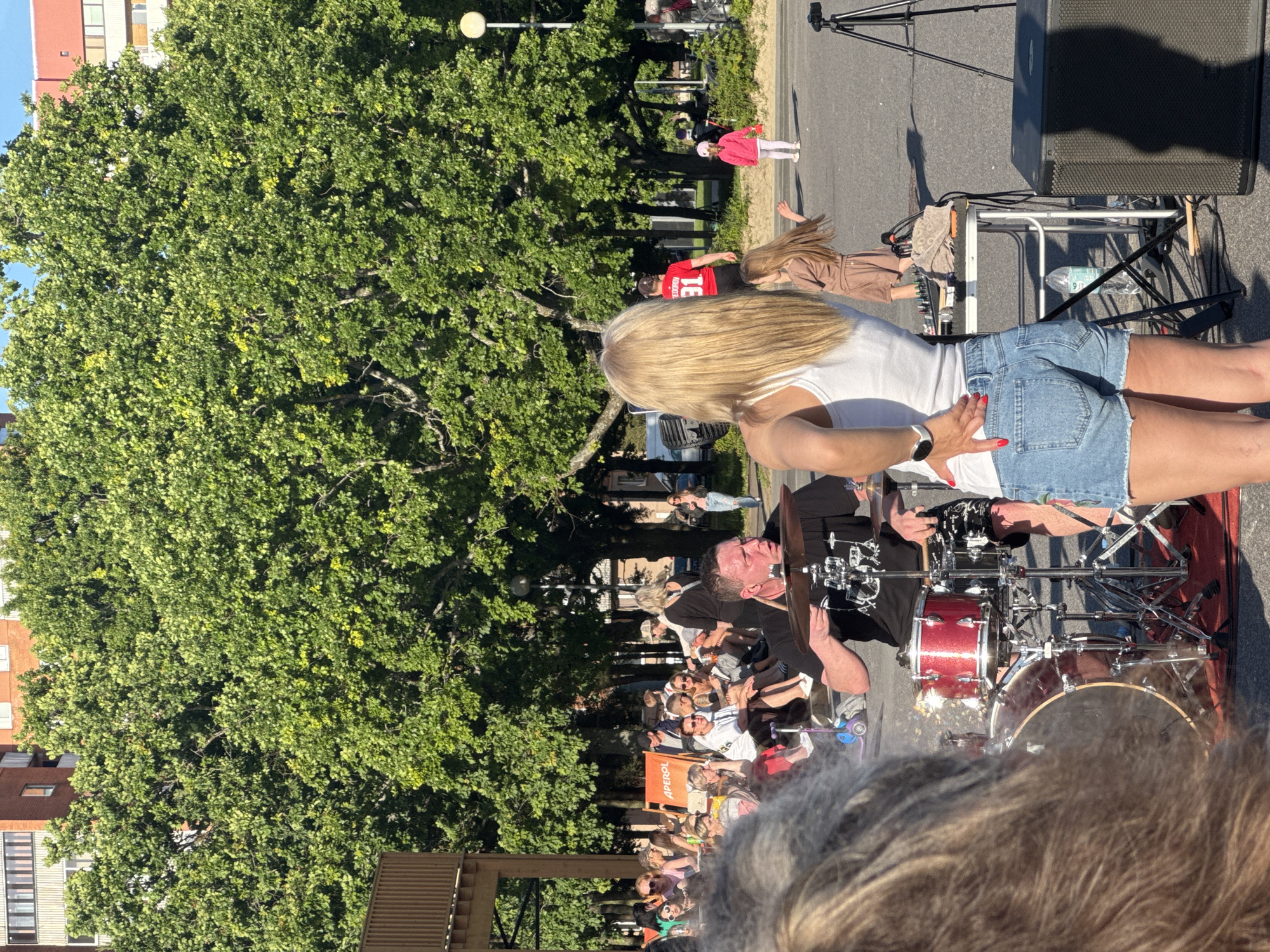 An outdoor daytime scene in Tallinn, Estonia, features a street performance with a drummer playing a red drum kit. He wears a black t-shirt and patterned shorts. A woman with blonde hair, wearing a white tank top and denim shorts, stands facing away from the camera, her hand on her hip. In the background, a crowd of people are seated on chairs and lounging on a deck chair. Large trees with green foliage are prominent, with apartment buildings visible behind them. A small child in a pink outfit is also in the background. A portable stage with equipment including speakers and a tripod is set up.