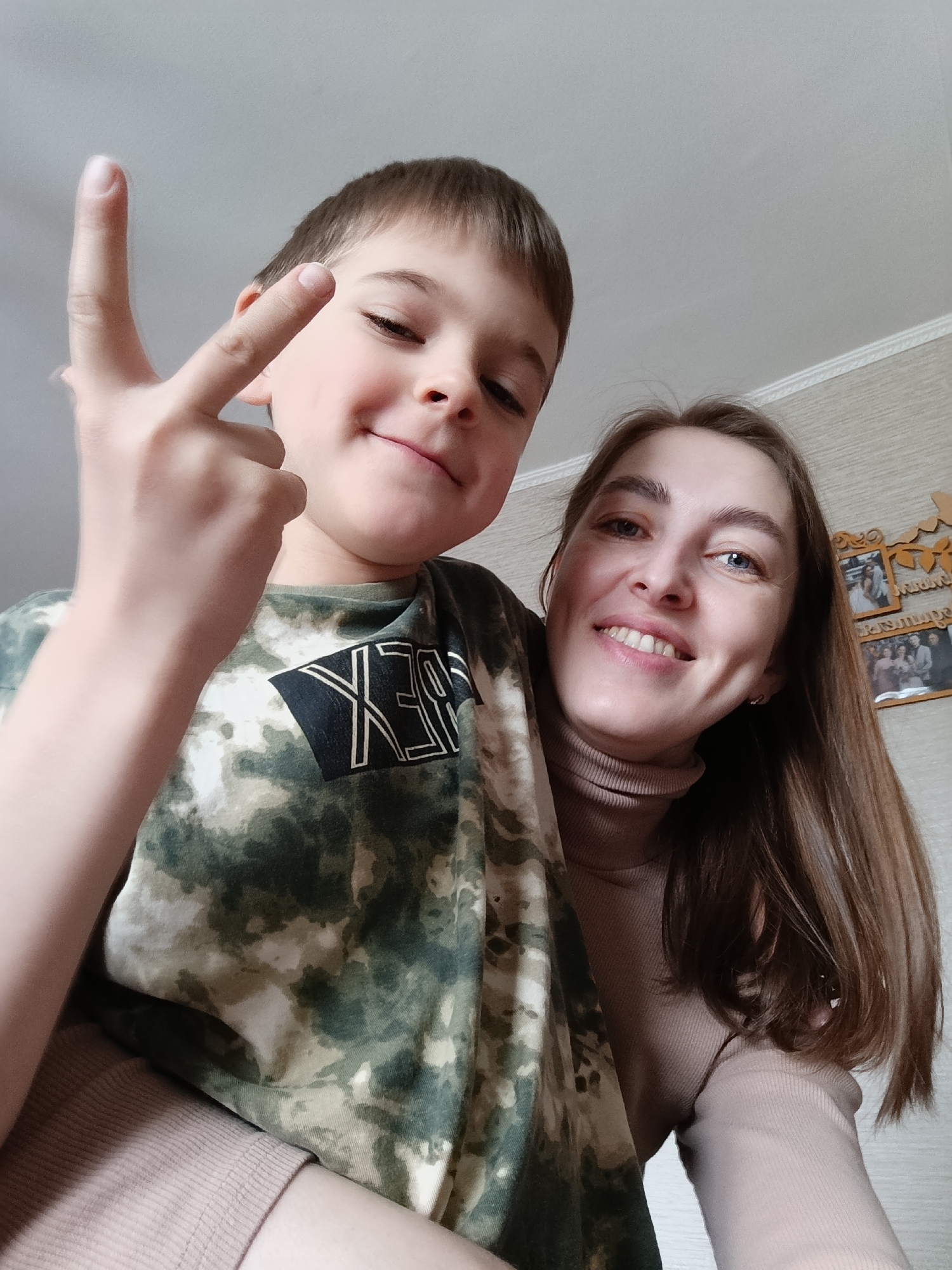 A boy and a woman are posing for a selfie. The boy is on the left and is wearing a camouflage t-shirt, and he is holding up a finger. The woman is on the right and is wearing a turtleneck sweater. They are both smiling. There are pictures hanging on the wall. This scene is taking place in the city of Фоки, Russia.