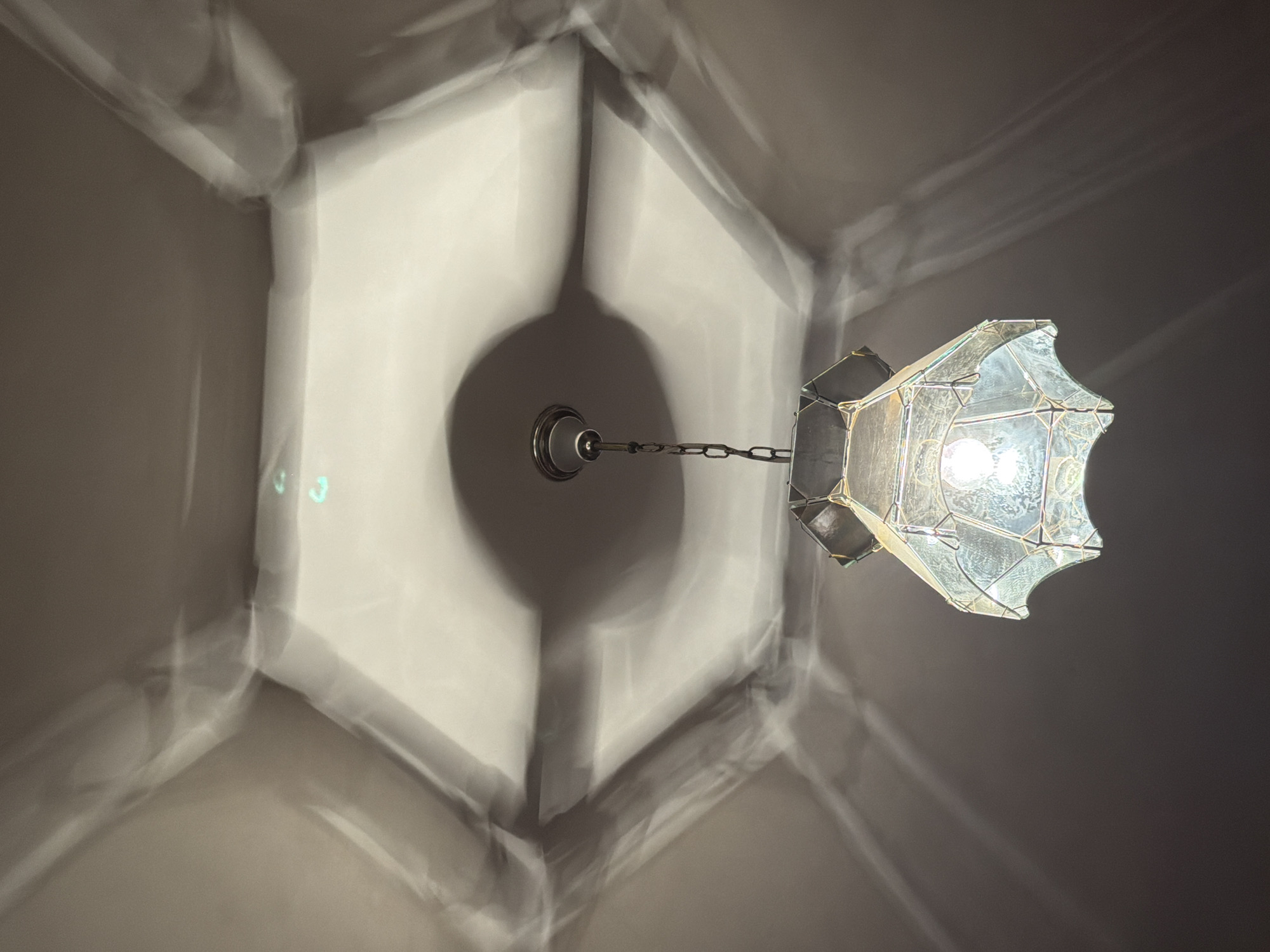 An antique chandelier hangs from a ceiling, casting intricate shadows. The ceiling has a hexagonal recess with a light fixture at its center. The chandelier itself is made of glass panels held together by a metal frame, with a single light bulb illuminating the space. The shadows projected onto the ceiling are a prominent feature of the image, creating a dramatic and somewhat abstract pattern. The overall scene is dimly lit, with the chandelier being the primary light source. The location is specified as Krumovgrad, Bulgaria.