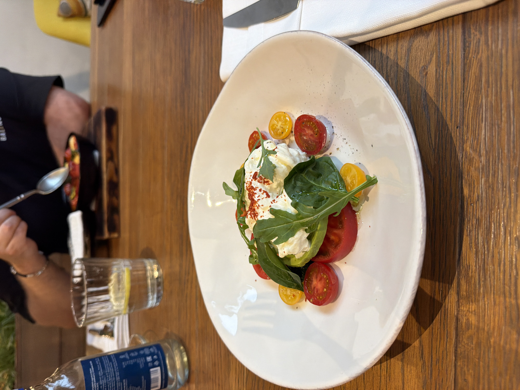 A photograph depicts a meal being eaten at a table. The table is wooden. A white plate is prominently displayed with a salad consisting of burrata, sliced tomatoes in red and yellow varieties, and green leafy vegetables. A knife rests on a white napkin near the plate. To the left of the plate are a water glass with a lemon slice, and a partially visible water bottle. A person, wearing a dark shirt, is seen holding a spoon and appears to be eating from a black bowl in the background. The context suggests an indoor restaurant setting. The location is not identifiable as Moscow, Russia, based on visual information.
