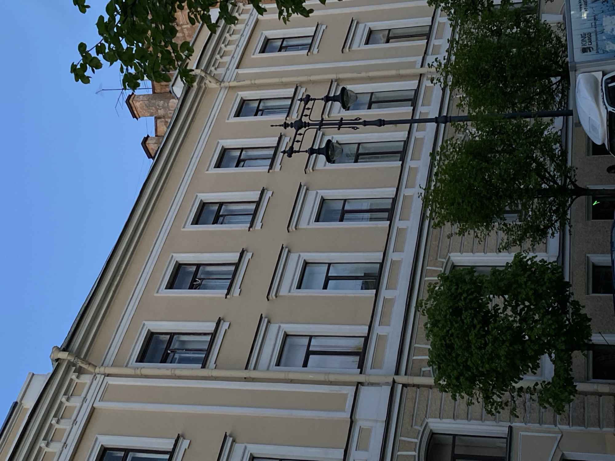 The photograph presents a building facade in Saint Petersburg, Russia. The architectural style incorporates symmetrical window placements with decorative white framing against a beige exterior. A black lamppost with two lights runs parallel to the building. Trees partially obscure the view of the building. The sky is visible in the upper-left corner.