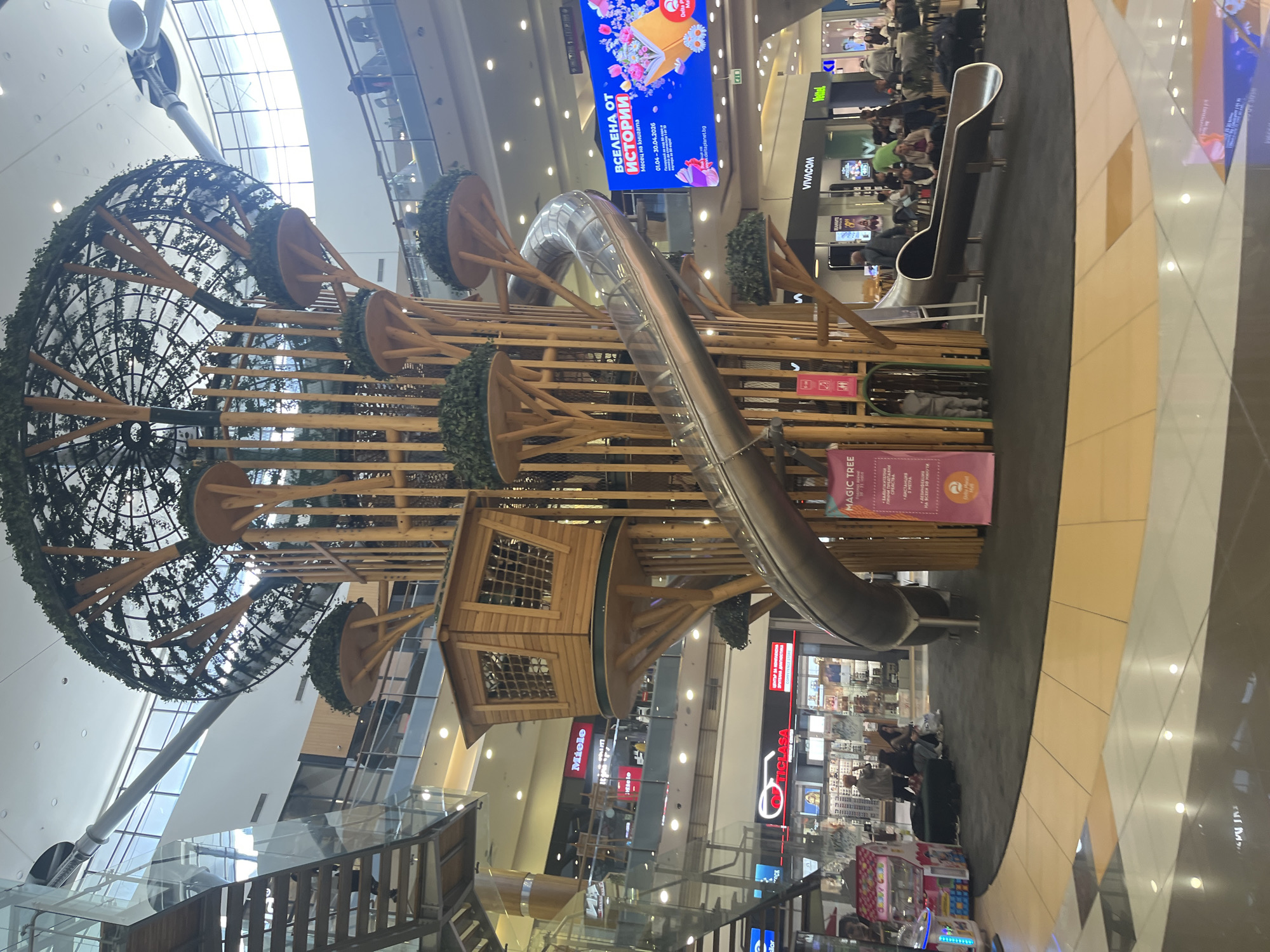 The image depicts a large, multi-level indoor playground structure inside a shopping mall in Unknown, Unknown. The playground is constructed primarily of wood and features a long, curving metal slide. The structure has multiple platforms resembling treehouses, and some are adorned with artificial foliage. The playground is decorated with several black, dome-shaped frames. The structure is positioned on a large black surface, and the surrounding mall area has shiny, tiled floors.
Several stores and advertisements are visible in the background. A large digital screen displays promotional content. People are present in the mall, some visible near the playground and others further back in the shopping area. A set of glass steps or a walkway is situated in the foreground, leading towards the playground structure. An arcade is in the bottom right.
