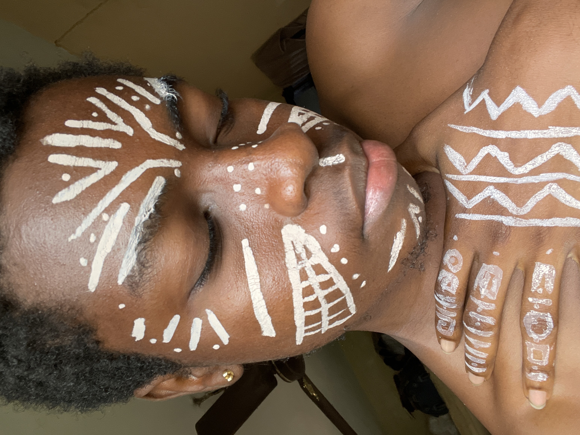 This close-up portrait, set in Nkpor, Nigeria, features a person with dark skin adorned with elaborate white body and face paint, depicted from the chest up. The subject's head is tilted, with eyes closed and lips slightly parted.
The white paint covers extensive areas of the face, including the forehead, temples, cheeks, nose, and chin, forming various intricate patterns. These designs consist of bold lines, some straight, others curved or zig-zagged, interspersed with numerous small dots. A distinct geometric pattern resembling a ladder or pyramid shape is visible on the cheek below the right eye. The paint also extends onto the visible hand and fingers, where similar linear and zig-zag motifs are present.
Notable details include the subject's short, dark, curly hair framing the face, and a small gold earring visible on the ear. The eyes are closed, lending a serene or contemplative expression, and the lips have a natural, light pink hue.
The visible text on the fingers of the hand reads "GOD IS" across the index and middle fingers, and "GOOD" across the ring and pinky fingers.
The setting appears to be indoors, with a warm, muted background in shades of brown and beige, suggesting ambient or controlled lighting rather than bright daylight. The overall tone of the image is artistic and culturally expressive, emphasizing the detailed body art.