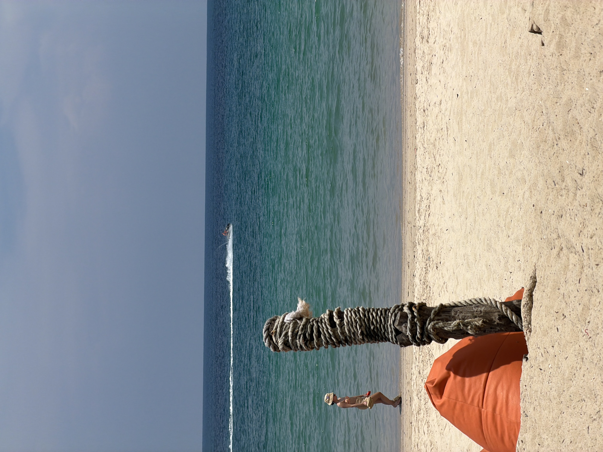 This image depicts a sunny beach scene in Choeng Thale, Thailand. The view is primarily split between the vast, turquoise-blue ocean on the left and the light tan sandy beach on the right, with a clear, light blue sky above the water.
In the foreground, a weathered wooden post, heavily wrapped with thick, dark rope, stands vertically in the sand. Perched calmly atop this post is a small, fluffy white cat, facing towards the ocean. To the right of the post, a vibrant orange bean bag chair is partially visible on the sand.
Further towards the water's edge, a person stands on the sand, facing the ocean. They are wearing light-colored shorts and a hat, possibly observing the activity or enjoying the view. In the distance on the ocean, a jet ski is actively moving, leaving a long, distinct white wake behind it on the calm water.
The scene suggests a warm, clear day with gentle ripples on the water. The beach appears clean and uncrowded, conveying a relaxed and serene atmosphere typical of a tropical resort area. There is no visible text in the image.