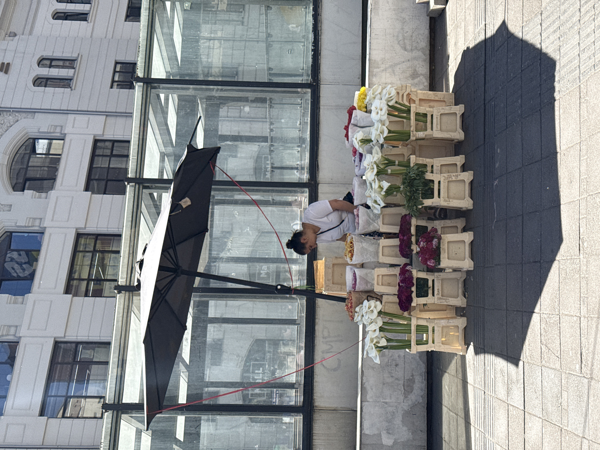 The image shows a woman selling flowers from a small flower stand on a sidewalk in Vracar Urban Municipality, Serbia. The flower stand is made up of several small, white wooden crates stacked on top of each other. The crates are filled with various types of flowers, including white lilies and purple flowers. The woman is wearing a white shirt and has her hair pulled back. She is standing behind the flower stand, tending to the flowers. An open black umbrella provides shade to the flower stand and vendor. Behind the woman is a glass-walled building with a white building behind it. The sidewalk is paved with grey rectangular blocks. The shadow of a building is visible on the sidewalk.