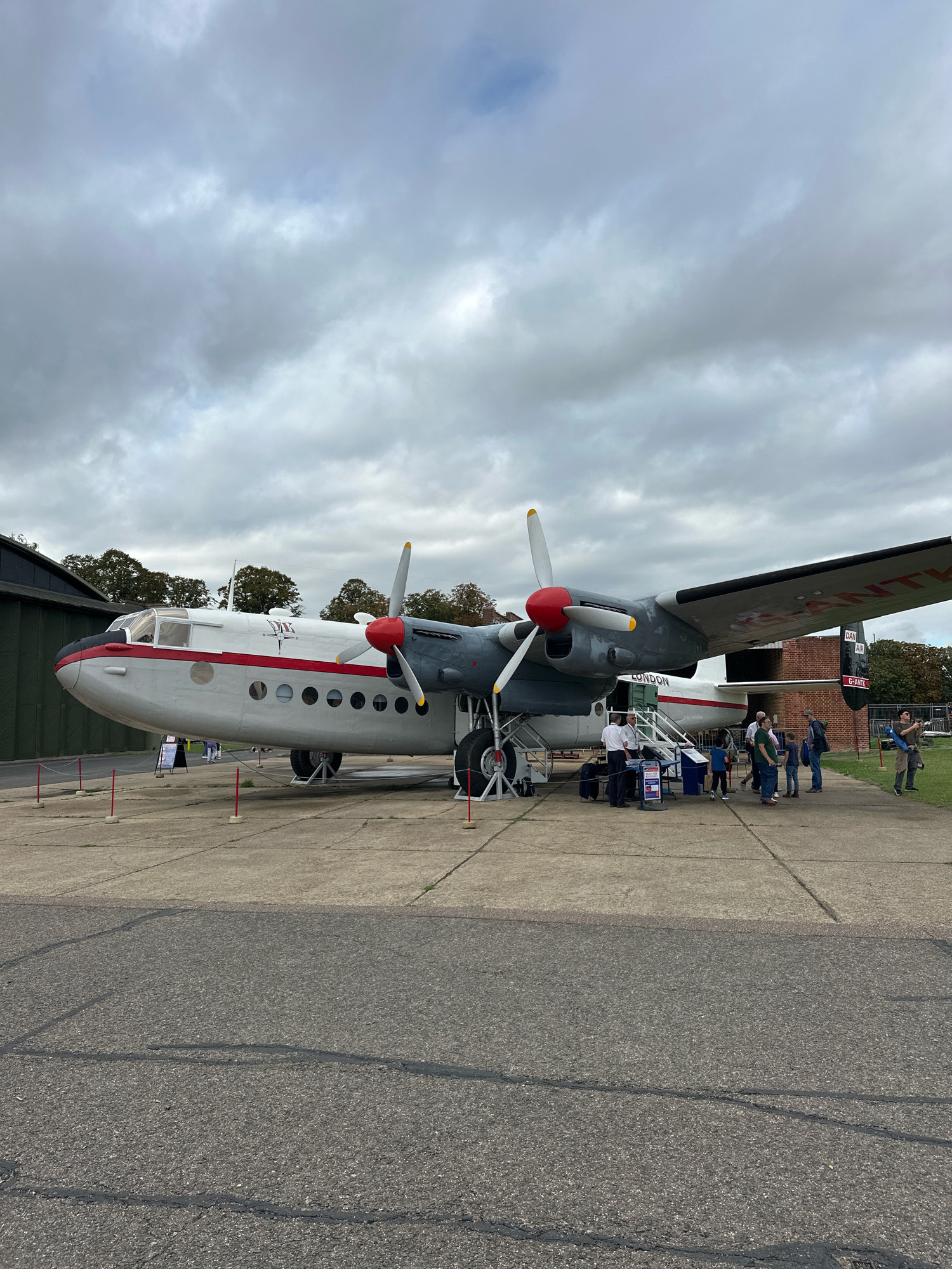 The image depicts a vintage twin-engine propeller aircraft on display outdoors, likely at an aviation museum or heritage site in Cambridge, United Kingdom. The aircraft is predominantly white with a red stripe running along its fuselage and red engine nacelles. It has large, grey wings with visible propellers and "DAN AIR LONDON" text visible on the tail. Several people, including adults and children, are gathered around the aircraft, some appearing to be boarding or observing it. Red stanchions with rope barriers are placed around the aircraft, suggesting it is an exhibit. In the background, a large, dark green hangar-like structure is visible, along with trees and a brick building. The ground is paved with concrete and asphalt, and the sky is overcast with grey clouds.