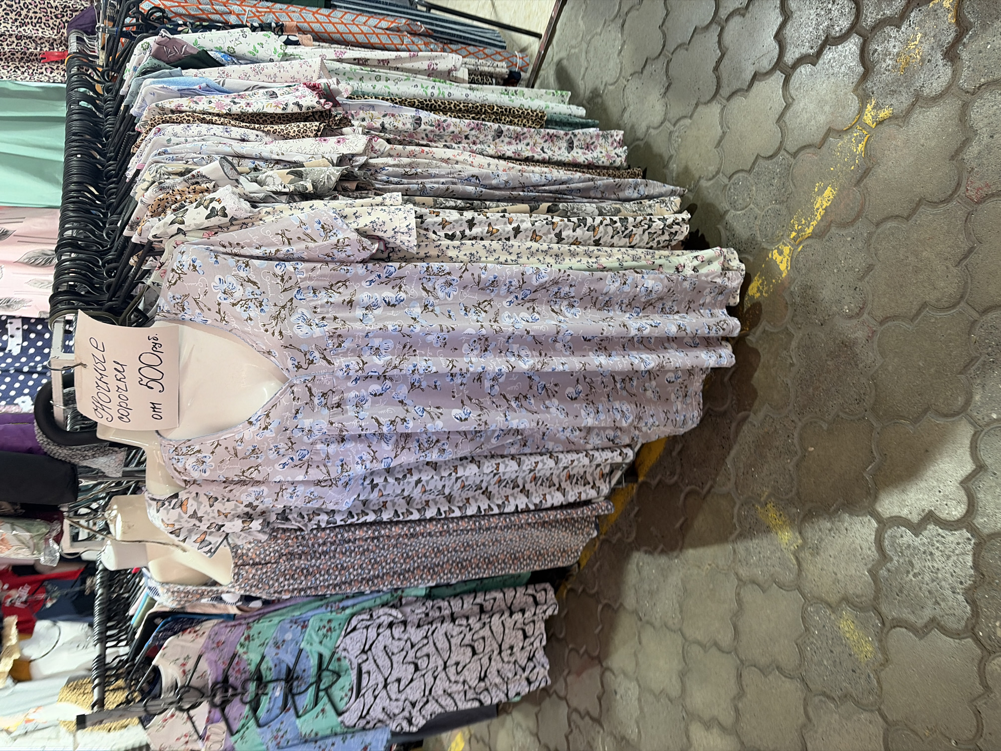 The image displays a retail stall or shop section featuring women's blouses or shirts, likely in a market setting in Sochi, Russia. The perspective is from slightly above, looking down a row of clothing.
In the foreground, a white mannequin torso displays a light lavender blouse adorned with delicate blue and white floral patterns. Behind this mannequin, numerous other blouses are neatly folded and stacked on black plastic hangers along a clothing rack. These garments showcase a variety of patterns, including different floral motifs, butterfly prints, and some animal-inspired (leopard) and abstract designs, predominantly in light and pastel colors.
A white price sign, attached to the clothing rack, is visible. The Cyrillic text on the sign reads "НОВЫЕ сорочки от 500 руб.", translating to "NEW shirts (or blouses) from 500 rubles," indicating the price point for the displayed items.
The floor beneath the clothing rack is paved with grey, clover-leaf shaped interlocking concrete blocks. Faint yellow paint markings are visible on some of these pavers, suggesting a public, possibly semi-outdoor or covered market environment. The overall scene is well-lit, with soft shadows cast on the pavement. No people are visible, but the arrangement clearly signifies a commercial display for sale.