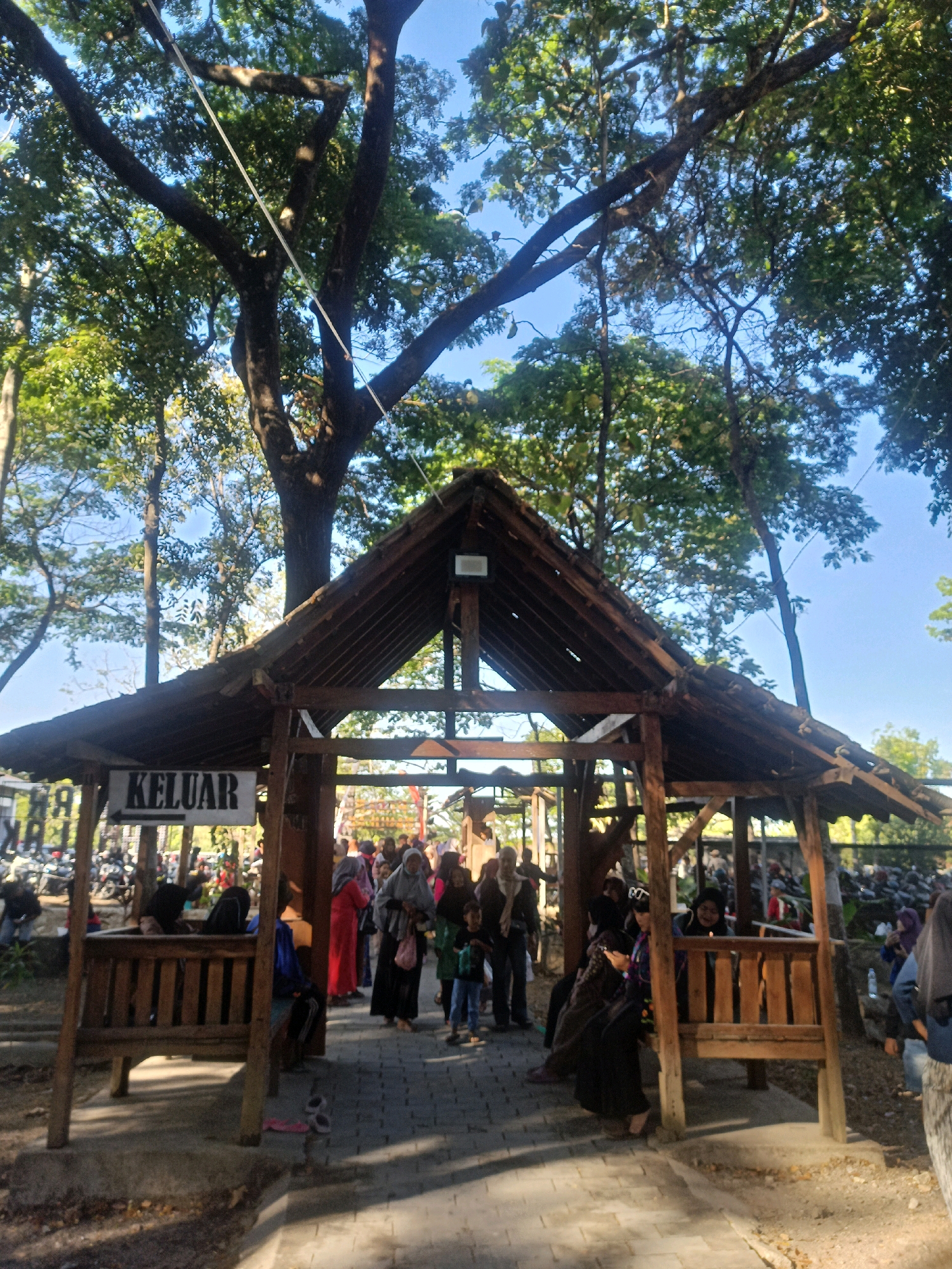 The image shows a wooden structure with a peaked roof, likely an entrance or exit point at an outdoor location. A sign with the word "KELUAR" in large black letters and an arrow pointing to the left is prominently displayed on the left side of the structure. The structure appears to be made of rough-hewn wood and has a rustic aesthetic.
A group of people are visible in and around the structure. Some are walking, while others are standing or sitting. They are dressed in casual clothing, with some individuals wearing headscarves. The environment appears to be a park or a public gathering space, with paved walkways and trees in the background. The presence of parked motorcycles on the left suggests it might be a tourist attraction or a venue where visitors arrive by motorcycle. The overall atmosphere seems to be one of activity and movement, possibly during a daytime event or a busy period. The background suggests a natural setting with lush greenery and a clear blue sky.