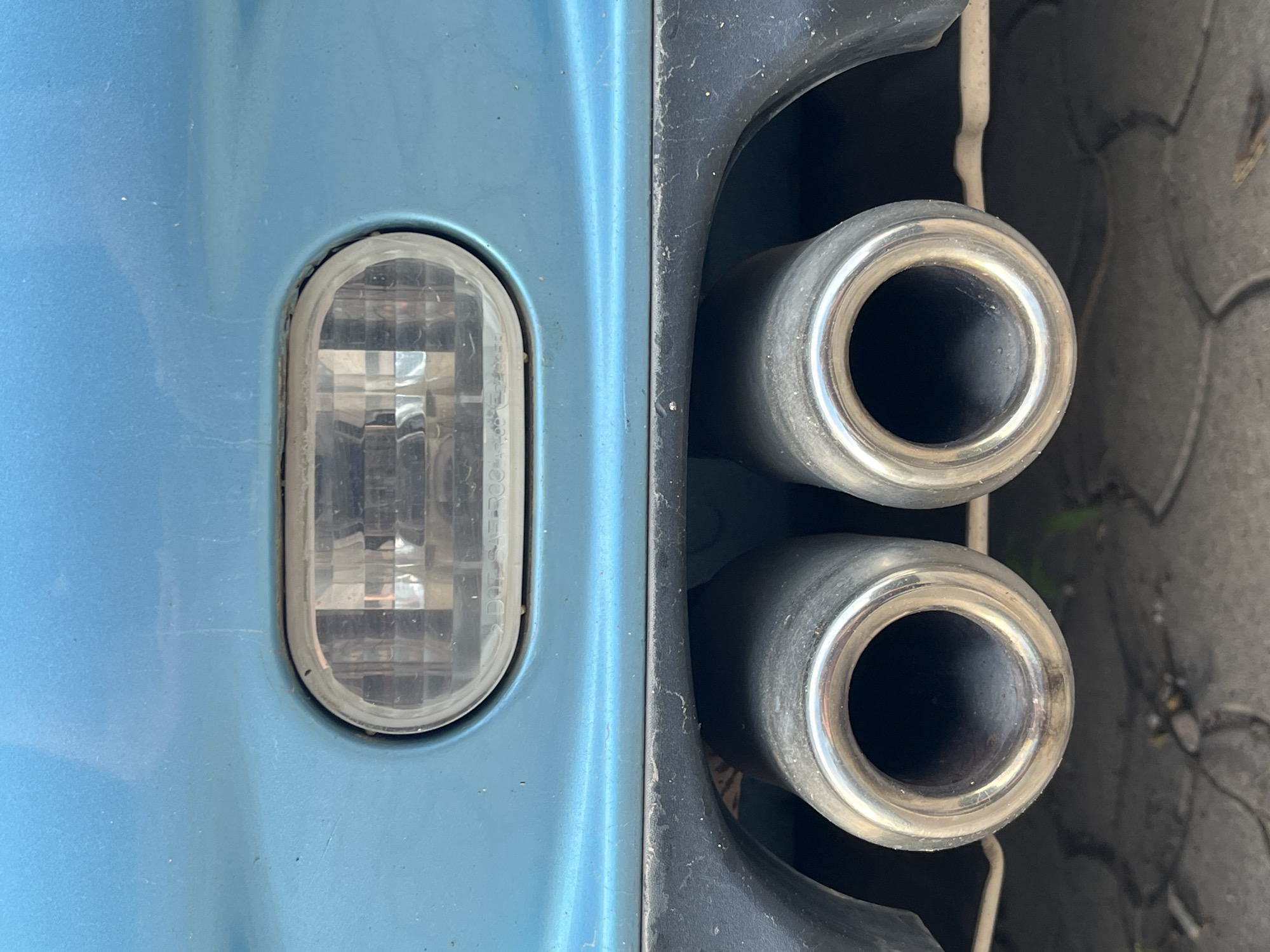 The image shows a close-up of the rear bumper and dual exhaust pipes of a blue car. Above the exhaust pipes, there is a clear, oval-shaped light fixture, likely a reflector or reverse light, embedded in the bumper. The car's bodywork is a bright blue, and the black plastic bumper trim is visible below the light. The exhaust pipes are made of polished metal, and their openings are visible. The background shows a paved surface, possibly a street or parking area, with some foliage visible at the bottom. The scene appears to be outdoors in daylight. There are no people or animals present in the image. The location is specified as Budva, Montenegro.