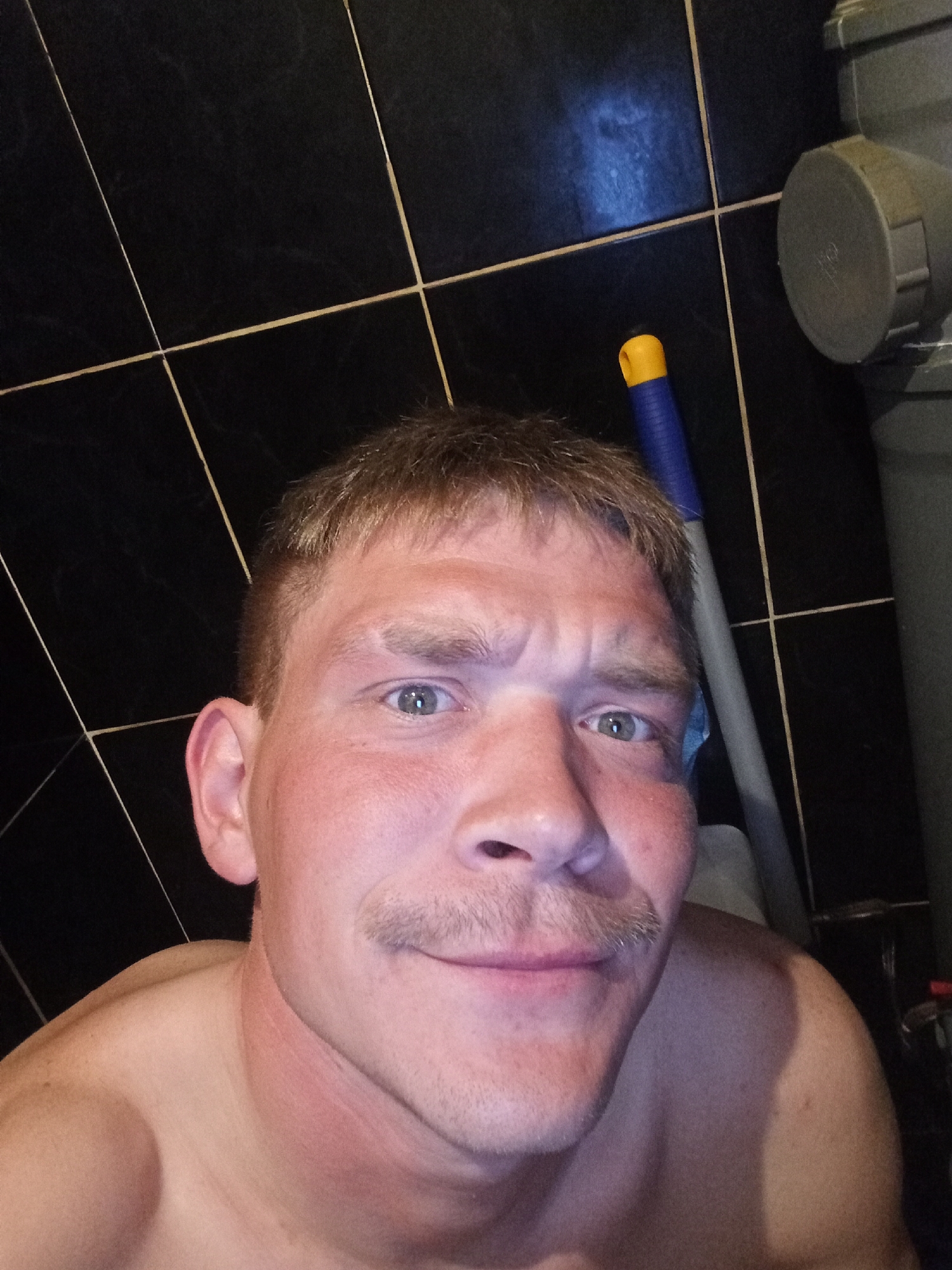 A fair-skinned man with short, light brown hair and a mustache is taking a selfie in a bathroom with black tiled walls. He is shirtless, and the top of his shoulder is visible in the lower left corner. The man is smiling slightly. In the background, a blue and yellow handled mop leans against the wall, and there is some sort of gray pipe fixture mounted on the wall to the right. The location is likely in a bathroom, though it is impossible to confirm whether it is in the city of Chaikovsky, Russia.
