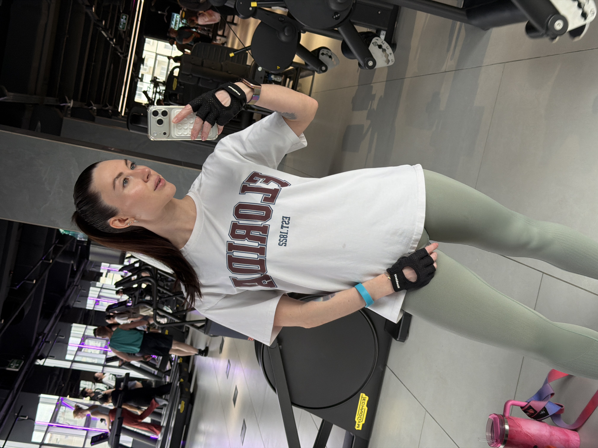 A woman is taking a mirror selfie in a gym. She's wearing a white T-shirt, green leggings, and gym gloves. The gym has treadmills and other exercise equipment. Other people can be seen working out in the background. The city or country can not be determined.