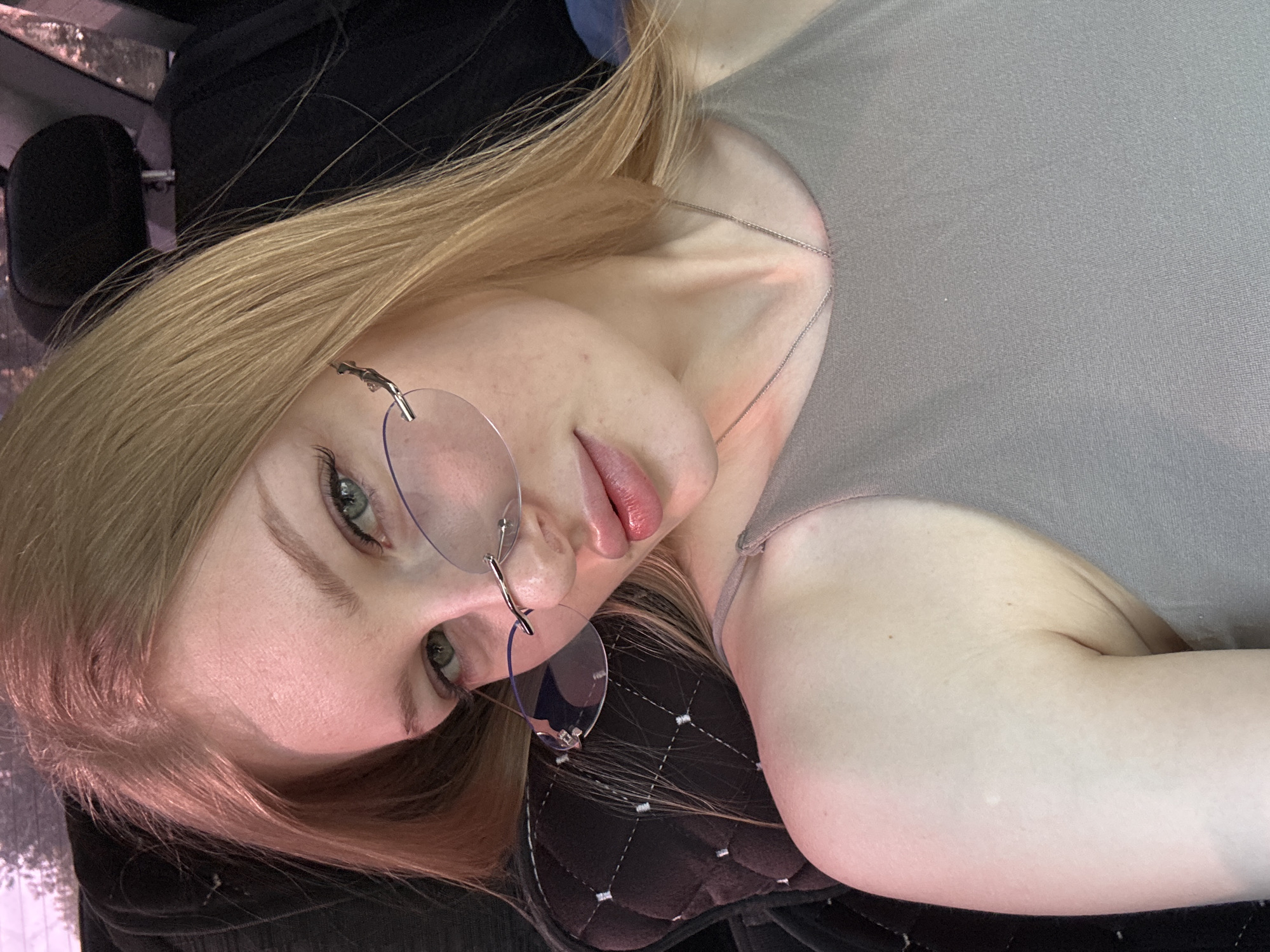 A young woman with blonde hair and light skin is lying down, wearing rimless glasses and a gray top. She has a silver necklace on. The background appears to be a car interior with black padded seats. No specific location in Perm, Russia, can be determined from the image.