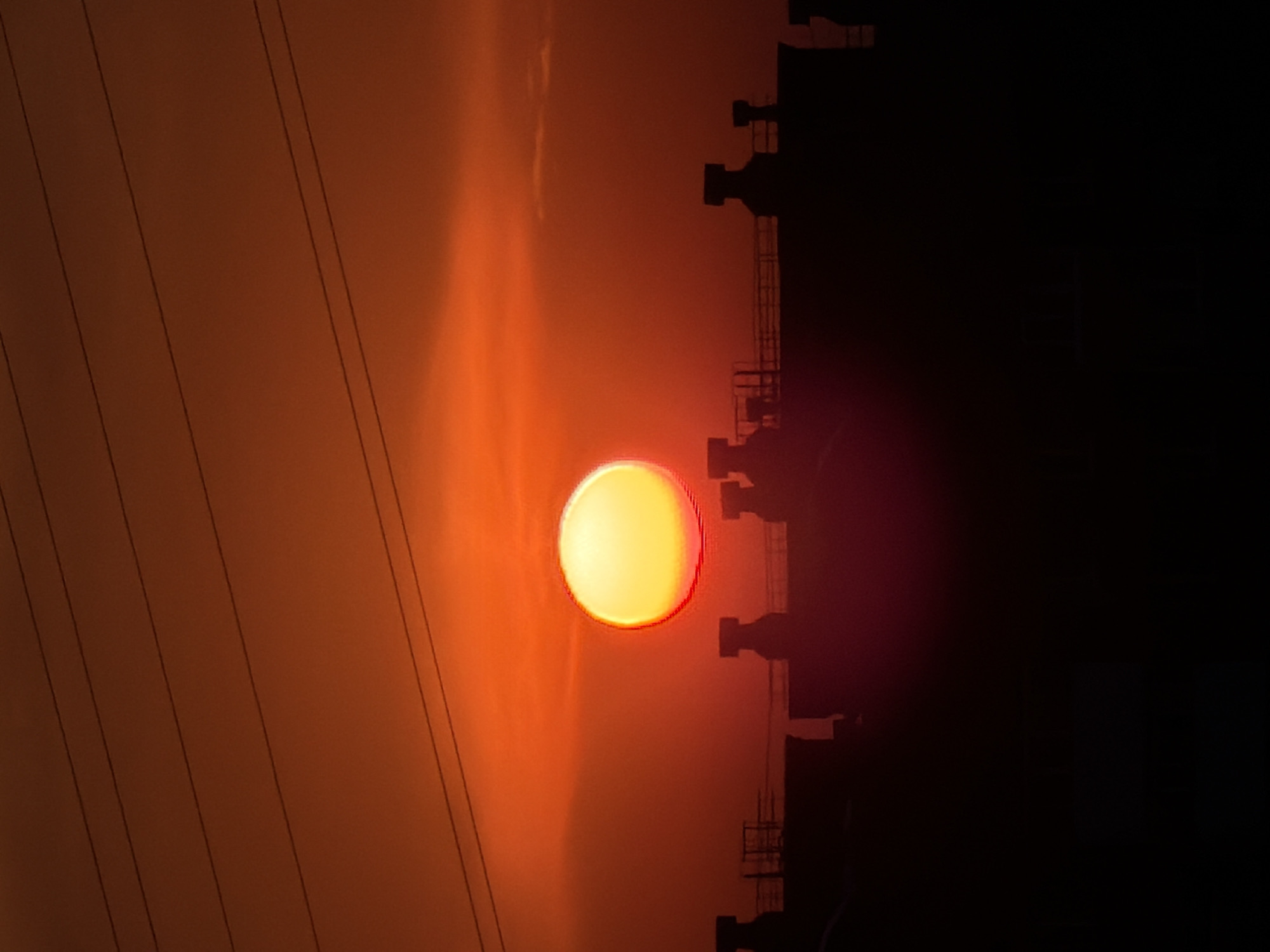 The image shows a sunset in Chaikovsky, Russia. The sun is setting behind a silhouette of industrial structures, possibly part of a factory or power plant. Power lines are visible in the foreground against the orange sky. The overall scene appears to be a blend of natural and industrial elements.