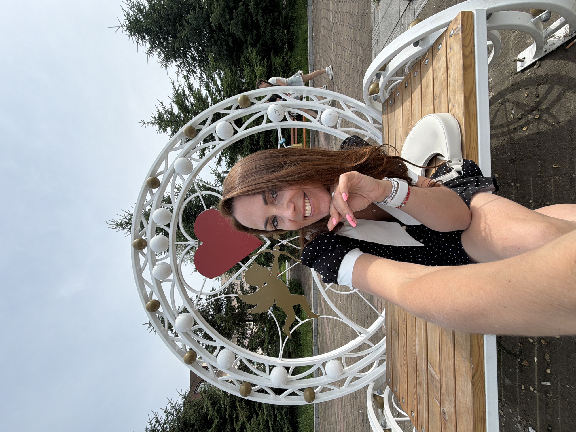 A medium shot captures a woman taking a selfie while sitting on a white, ornate bench with a wooden seat. The bench is situated outdoors in what appears to be a park or public square. Behind the woman is a large, white, decorative archway featuring a red heart and a golden cupid figure. The archway is adorned with white and gold spheres. Evergreen trees are visible in the background, and a paved walkway with brick patterns can be seen. A second woman, in a light-colored skirt and top, walks in the distance. The woman in the foreground has long, brown hair and is smiling at the camera, her hand resting on her chin. She is wearing a black and white polka dot dress and jewelry on her wrist. A white handbag rests on the bench beside her. The sky is overcast.