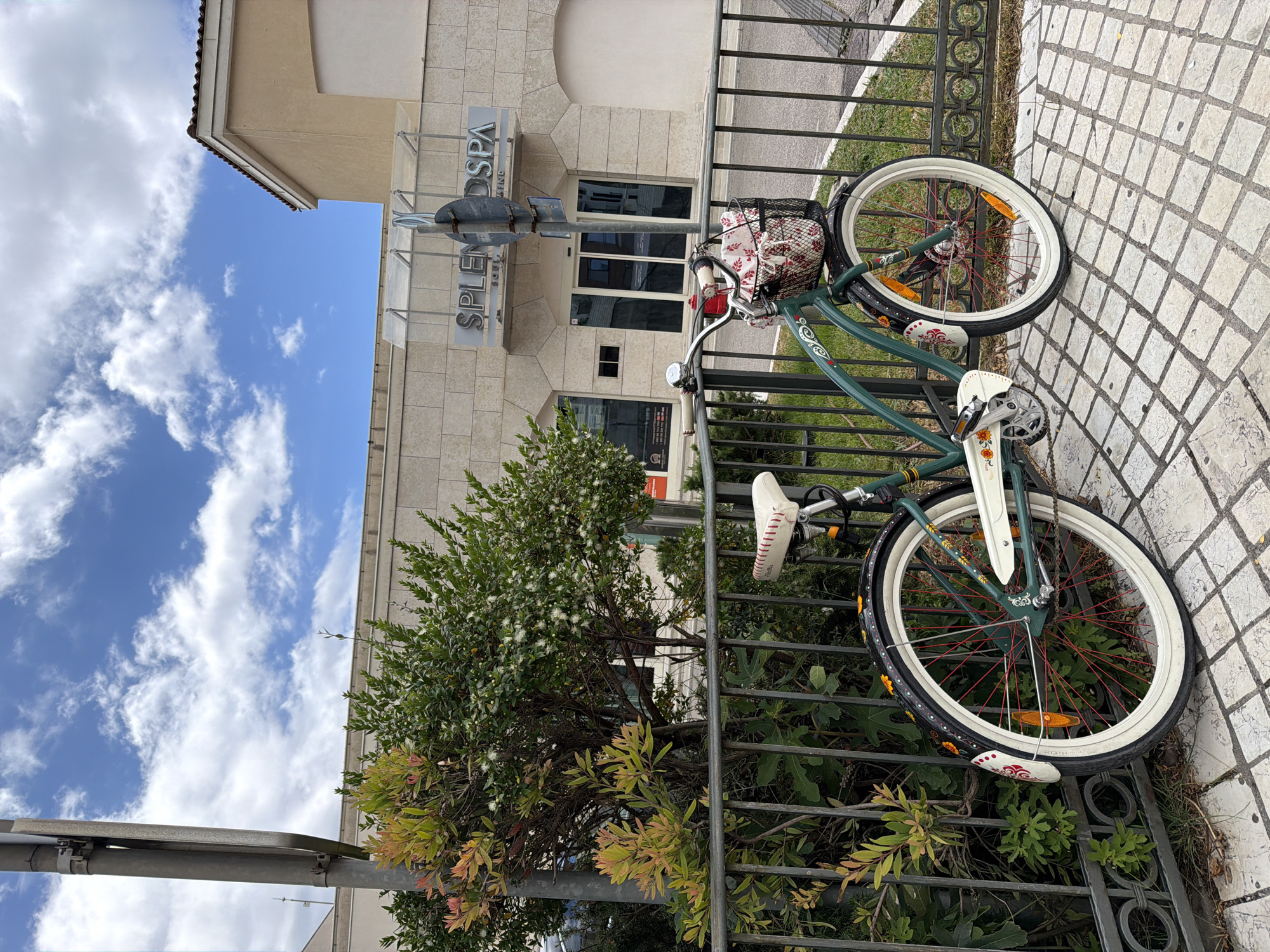 A vibrant green bicycle with white fenders and wide white-walled tires is parked on a light-colored, diamond-patterned tiled sidewalk. It leans against a dark wrought-iron fence that features decorative elements at its base. The bicycle's wheels have distinctive red spokes and orange reflectors, and its white seat is detailed with red stitching. Floral motifs decorate the fenders. A black wire basket on the handlebars contains a white and red patterned fabric.
Behind the fence, a dense, verdant bush with numerous small white flowers flourishes, partially obscuring the lower part of a modern building. This light-colored building, constructed from a tiled or stone material, features multiple dark glass doors and windows. A prominent silver sign on its facade clearly displays the text "SPLENDO SPA."
Above the building and foliage, a bright, clear blue sky is dotted with fluffy white cumulus clouds, indicating a sunny day, likely morning or afternoon. The scene captures a quiet, leisurely moment in Boreti, Montenegro.