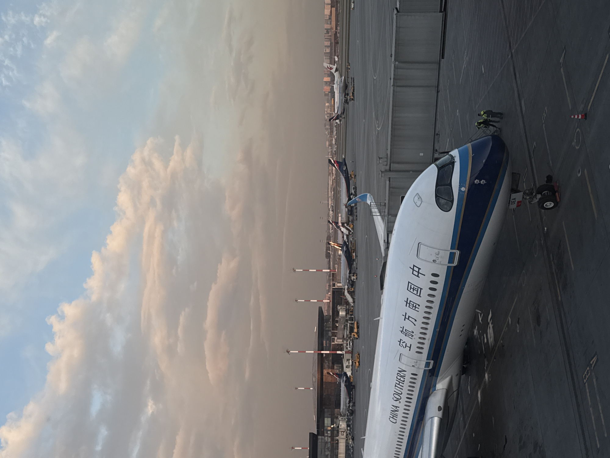 The image displays an active airport tarmac in Khimki, Russia, likely Sheremetyevo International Airport. A China Southern Airlines aircraft dominates the foreground, with its nose and front fuselage visible. The airline's name, "CHINA SOUTHERN," and its Chinese counterpart, "中国南方航空," are clearly legible in blue text on the white body of the plane. Below the text, a series of small, dark circles, possibly windows, runs along the fuselage.
Two ground crew personnel, dressed in high-visibility vests, are seen near the nose of the China Southern aircraft, engaged in tasks that appear to be pre-flight checks or ground handling procedures. A towbar is connected to the aircraft's nose wheel, and a piece of ground equipment with "DEA" partially visible is also present.
In the mid-ground and background, several other parked airplanes are visible on the tarmac, including one with a distinctive red tail logo, possibly indicating Japan Airlines. Airport buildings and numerous light poles with red and white stripes punctuate the distant horizon. The sky, which occupies the left portion of the image, features soft, diffused clouds against a light blue backdrop, tinged with warm orange and pink hues, suggesting either sunrise or sunset. The overall scene captures routine airport operations during a period of soft, atmospheric lighting.