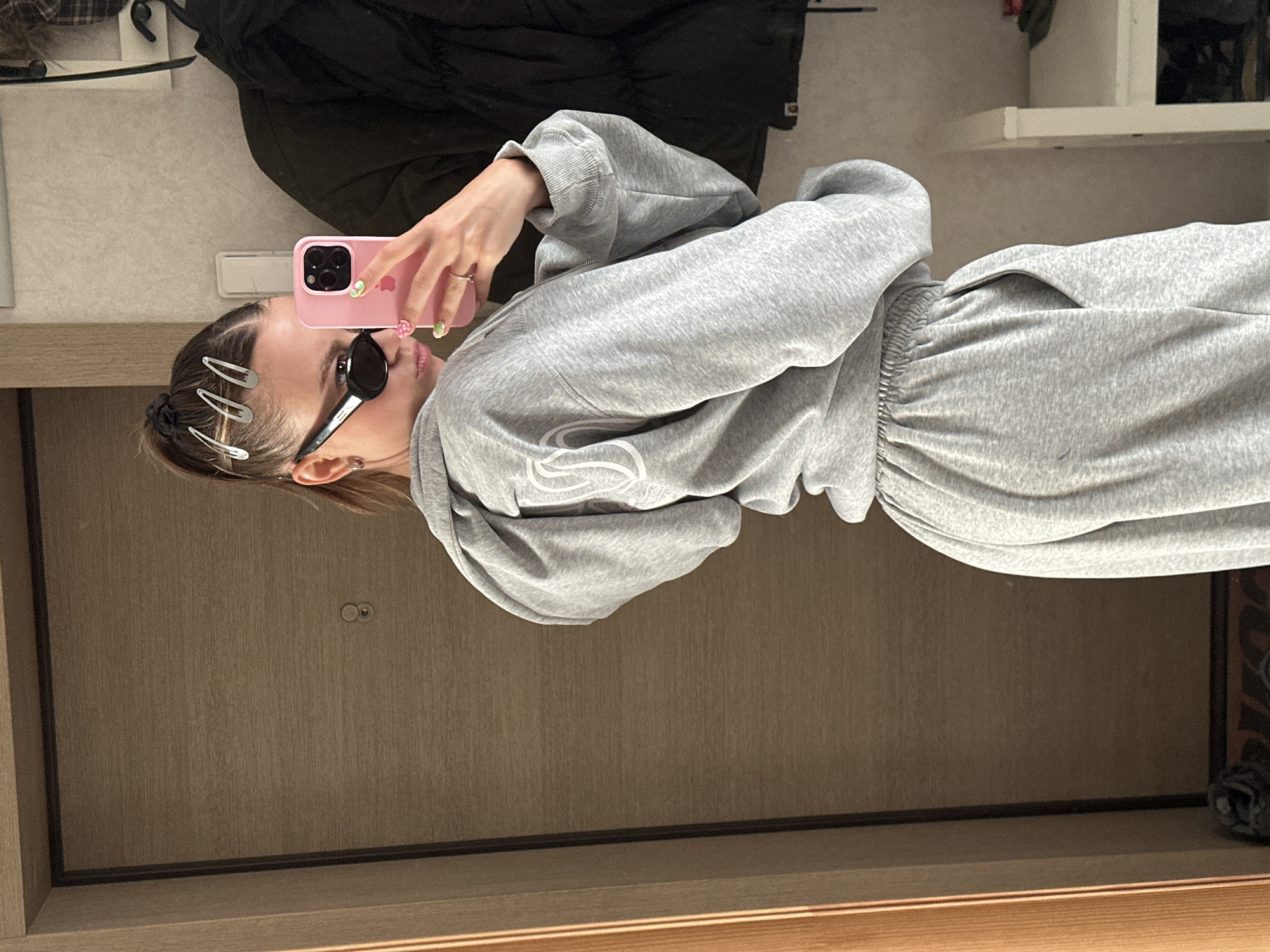 A young woman is taking a mirror selfie in what appears to be a hallway or entryway. She is wearing a grey tracksuit and black sunglasses, with several silver hair clips in her hair. She is holding a pink phone in her right hand. The background shows a wooden door on the left and a wall with a coat hanging on it to the right. The overall scene suggests an indoor domestic setting, likely in Moscow, Russia, given the style of the door and the general aesthetic.