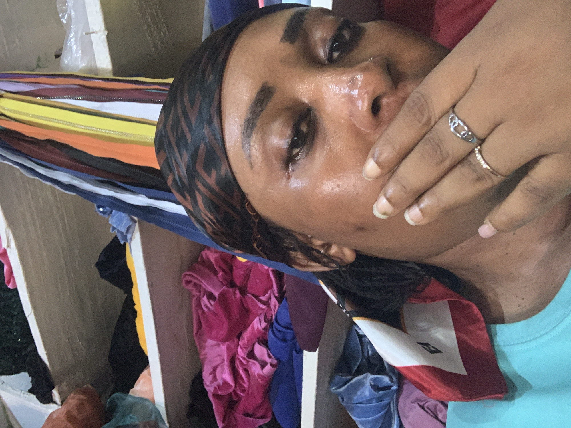 A person with dark skin and wet eyes is covering their mouth with their right hand. They are wearing a patterned headscarf and a light blue shirt. Two rings are visible on their hand. The person appears to be lying down or reclining. In the background, there are several zippers of varying colors and pieces of fabric in shades of pink, blue, and black. The setting appears to be indoors. Sabon Tasha, Nigeria is not verifiable based on the image.
