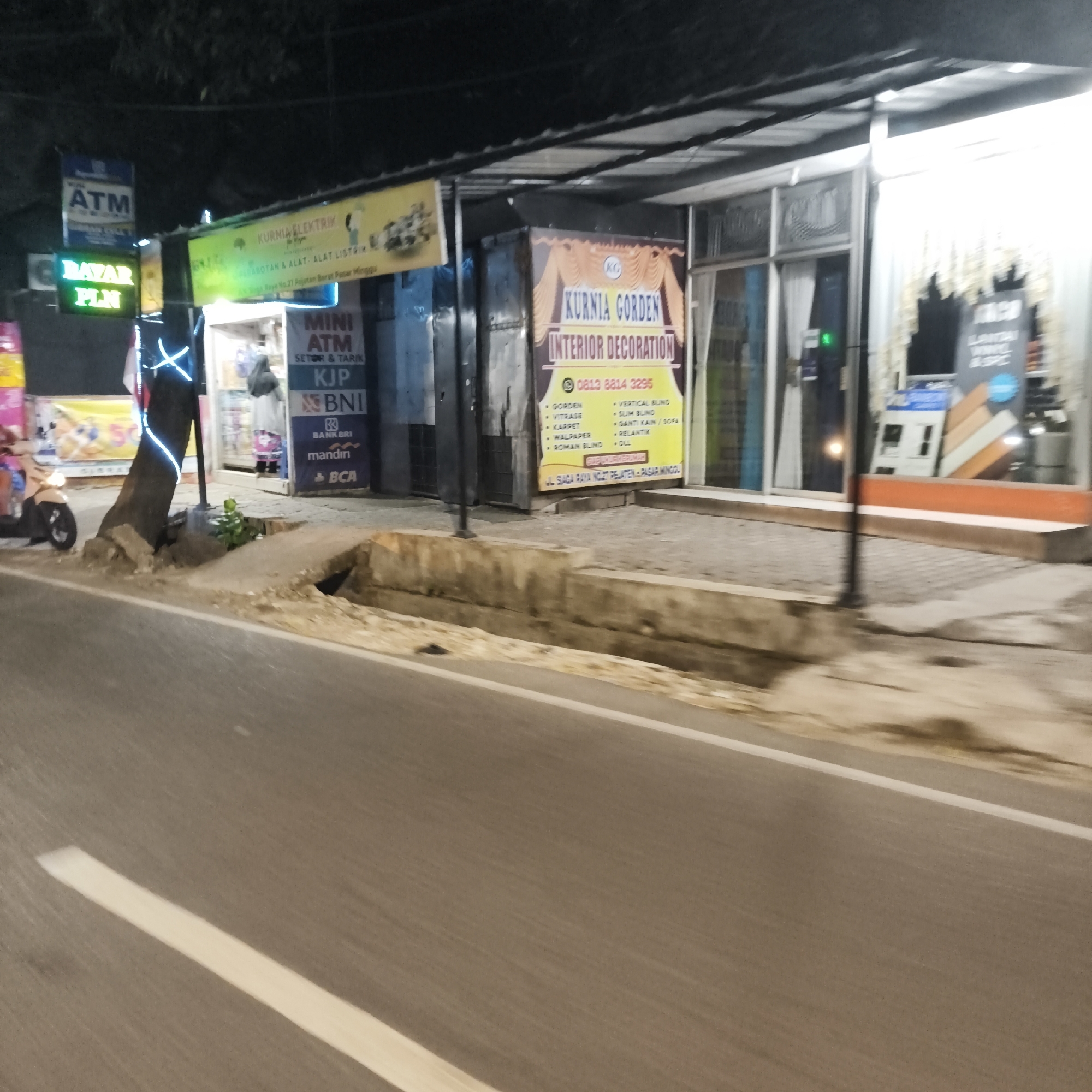 The image depicts a street scene at night in South Jakarta, Indonesia. The foreground shows a road with a white line marking the edge. In the mid-ground, there is a sidewalk with a broken concrete planter box. Along the sidewalk, there are several storefronts. One storefront has a yellow sign with "KURNIA GORDEN INTERIOR DECORATION" written on it, along with a phone number. Another storefront has multiple ATM signs, including "MINI ATM SETOR & TARIK KJP BNI mandiri BCA". There are also neon signs visible, including a green "BAYAR PLN" sign and a blue ATM sign. A tree wrapped in blue neon lights stands near the ATM storefront. A scooter is parked on the left side of the image. The background shows the dark night sky and the upper portions of buildings. The scene appears to be in a commercial area.