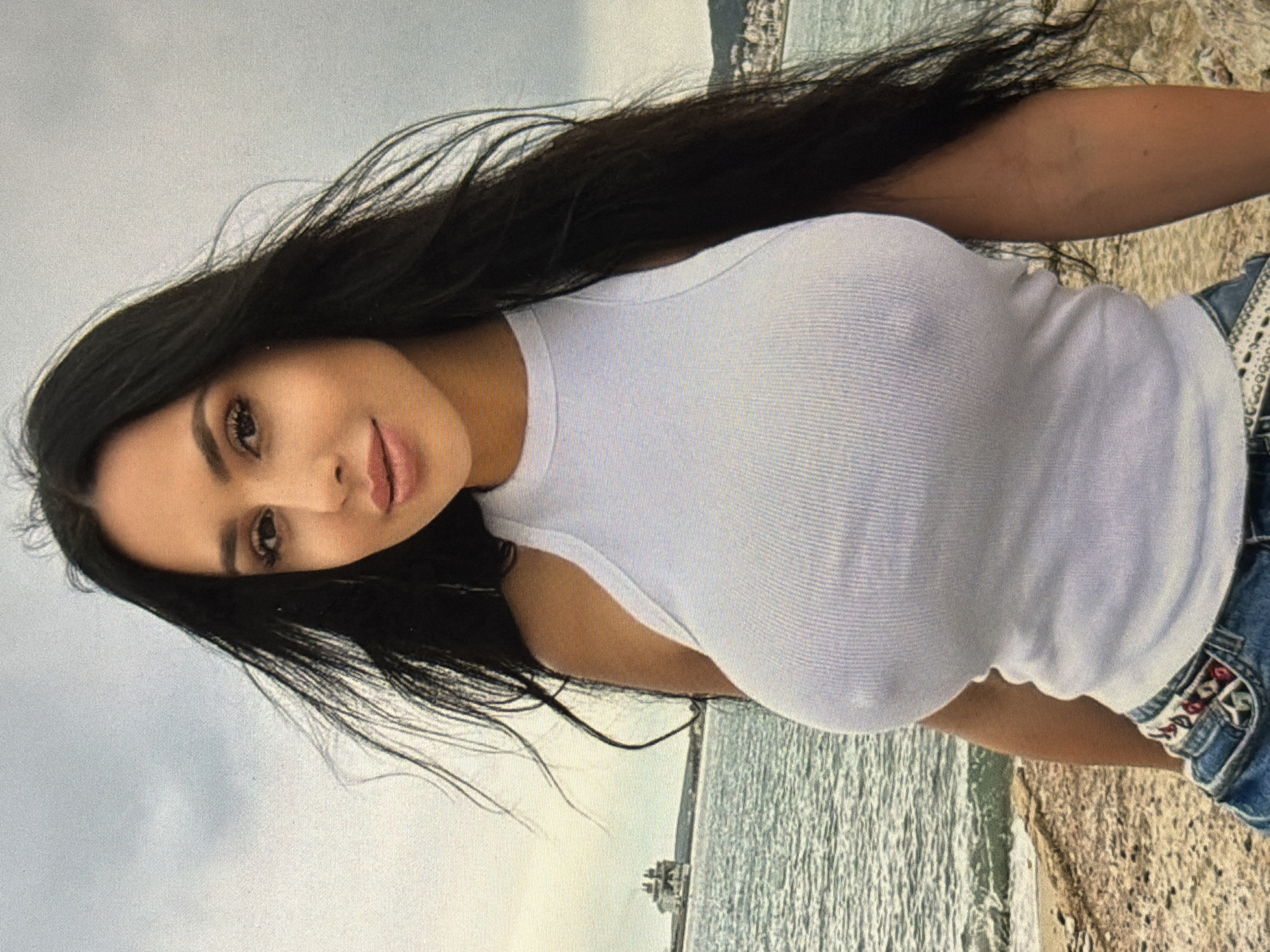 The image depicts a medium shot of a young woman with long, dark hair standing outdoors with a body of water and a cloudy sky in the background. She is wearing a white, ribbed tank top and denim shorts. The woman is looking towards the camera with a slight smile. The location is not specified as Shumen, Bulgaria, and there are no identifiable landmarks to suggest that location. The environment appears to be a coastal area with a beach and some rocky terrain. A ship is visible in the distance on the water.