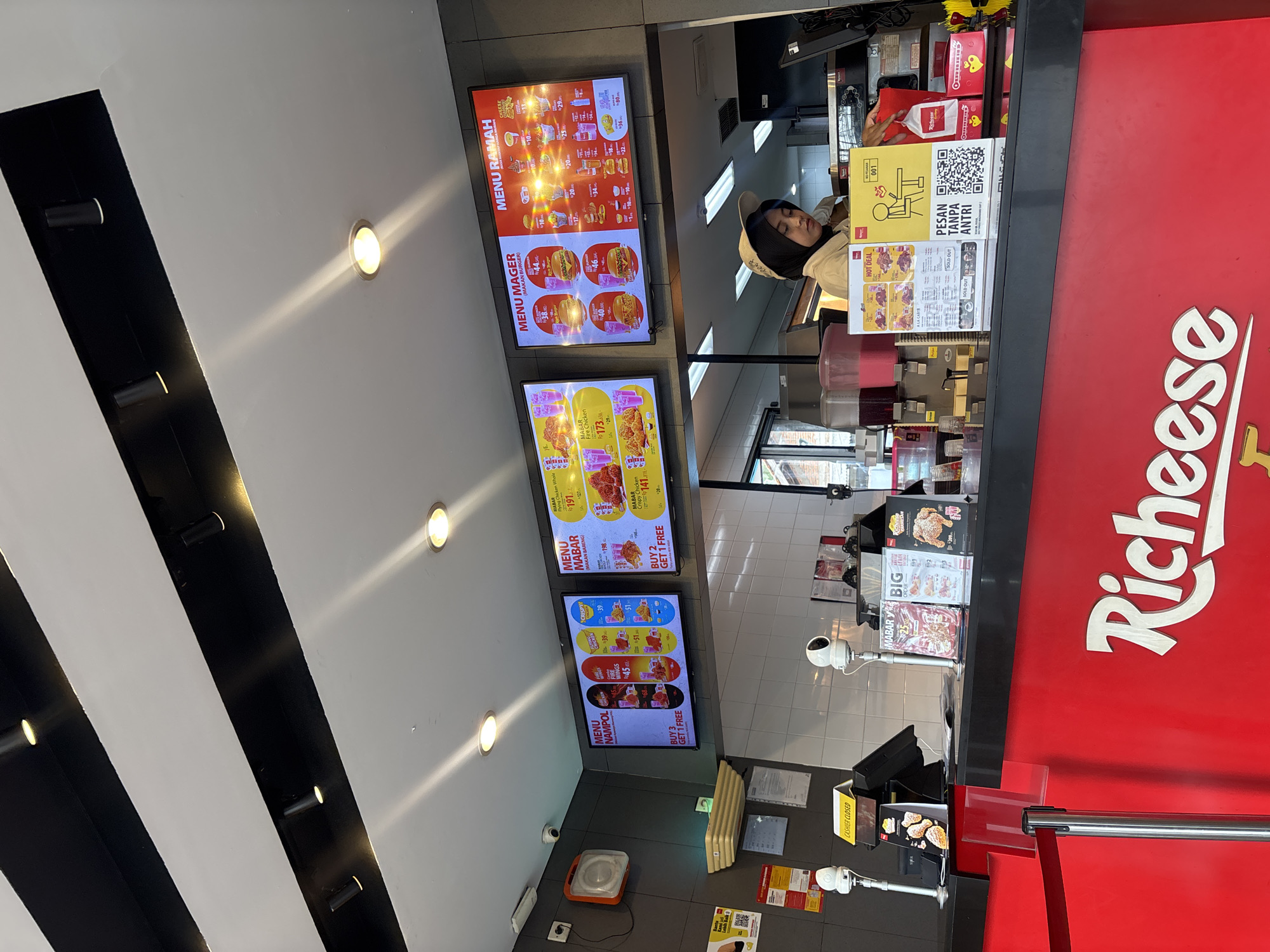 The image shows the interior of a Richeese Factory fast-food restaurant in Tangerang, Indonesia. The scene is captured from a low-angle perspective, looking towards a counter area. On the counter, there are several menu boards displaying food items and prices, including digital screens and printed posters. A female employee wearing a beige cap and hijab is visible behind the counter, attending to customers or preparing orders. Several security cameras and a customer queue management system are also present. The restaurant's prominent red branding is visible on the counter and on merchandise. The overall environment suggests a busy, operational fast-food establishment.