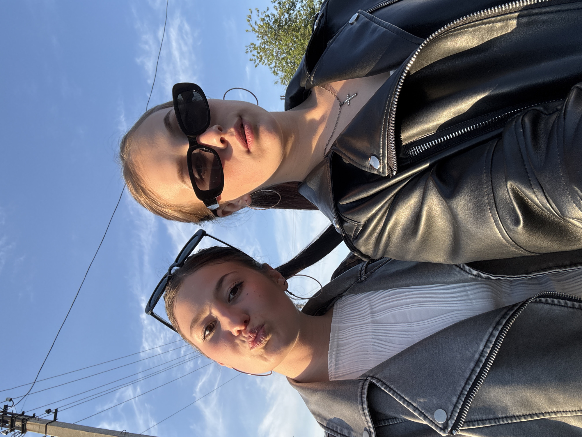 Two young women are posing for a selfie taken from a low angle. The woman on top is wearing a black leather jacket, a silver necklace with a cross pendant, and black sunglasses. The woman below is wearing a gray leather jacket, a white shirt, and her sunglasses are propped on her head. Both women have hoop earrings. The background consists of a bright blue sky with some wispy clouds. Utility poles and wires are visible in the lower left corner of the frame. Based on the limited visual information, there is not sufficient data to determine the exact location in the city of Фоки, Russia.