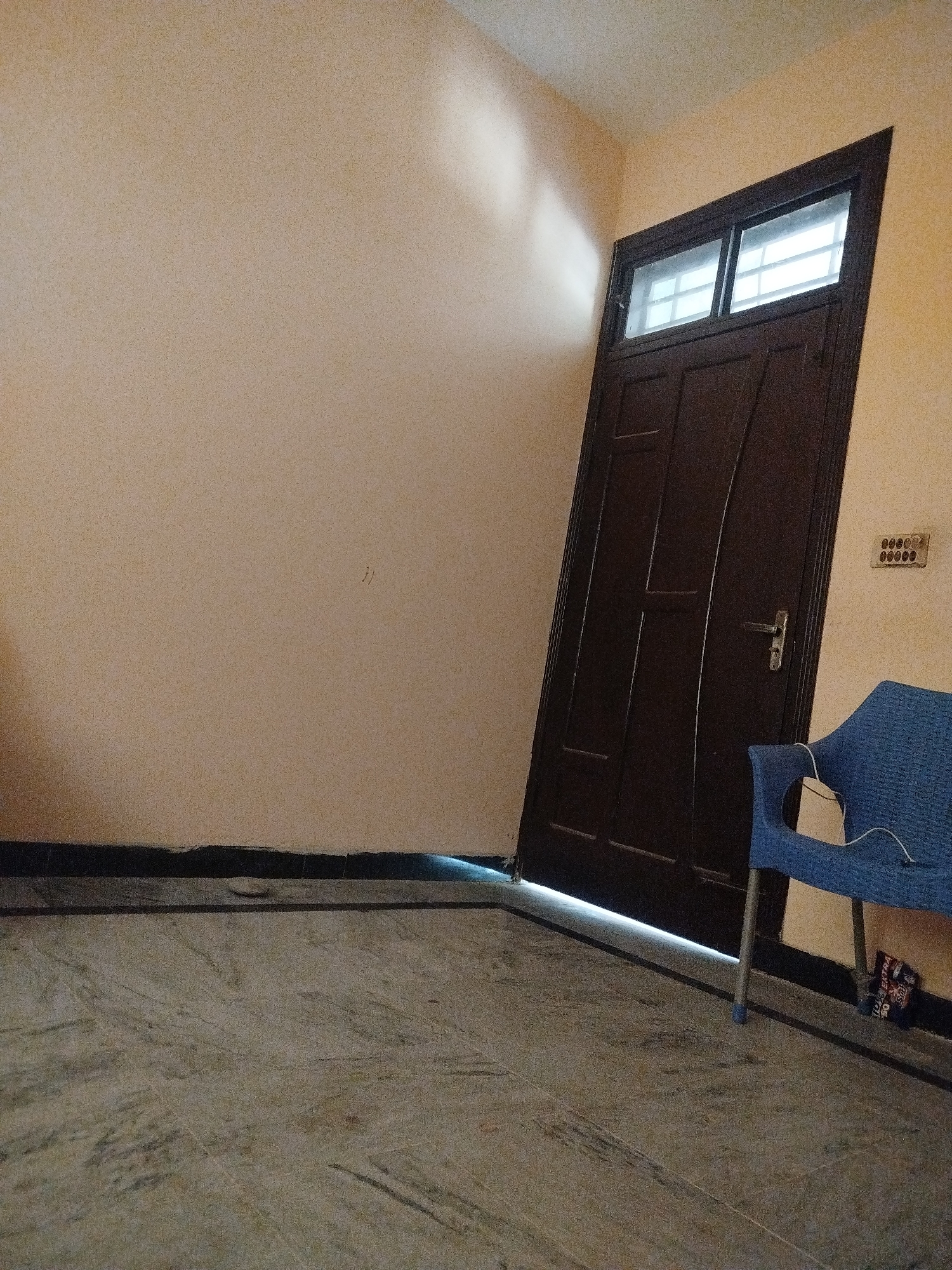 The image depicts a room with a light brown painted wall on the left and a darker beige painted wall on the right. A dark brown wooden door with two rectangular windows at the top is positioned diagonally across the room, leaning against the right wall. The door has a silver handle. To the right of the door, a blue plastic chair is placed with a white cord draped over it. A power outlet with multiple sockets is mounted on the wall above and to the right of the chair. The floor is tiled with a marble pattern in shades of gray and brown. A black baseboard runs along the bottom of the walls. The scene appears to be an interior space, possibly a room in a residential building, located in Lassan, Pakistan.
